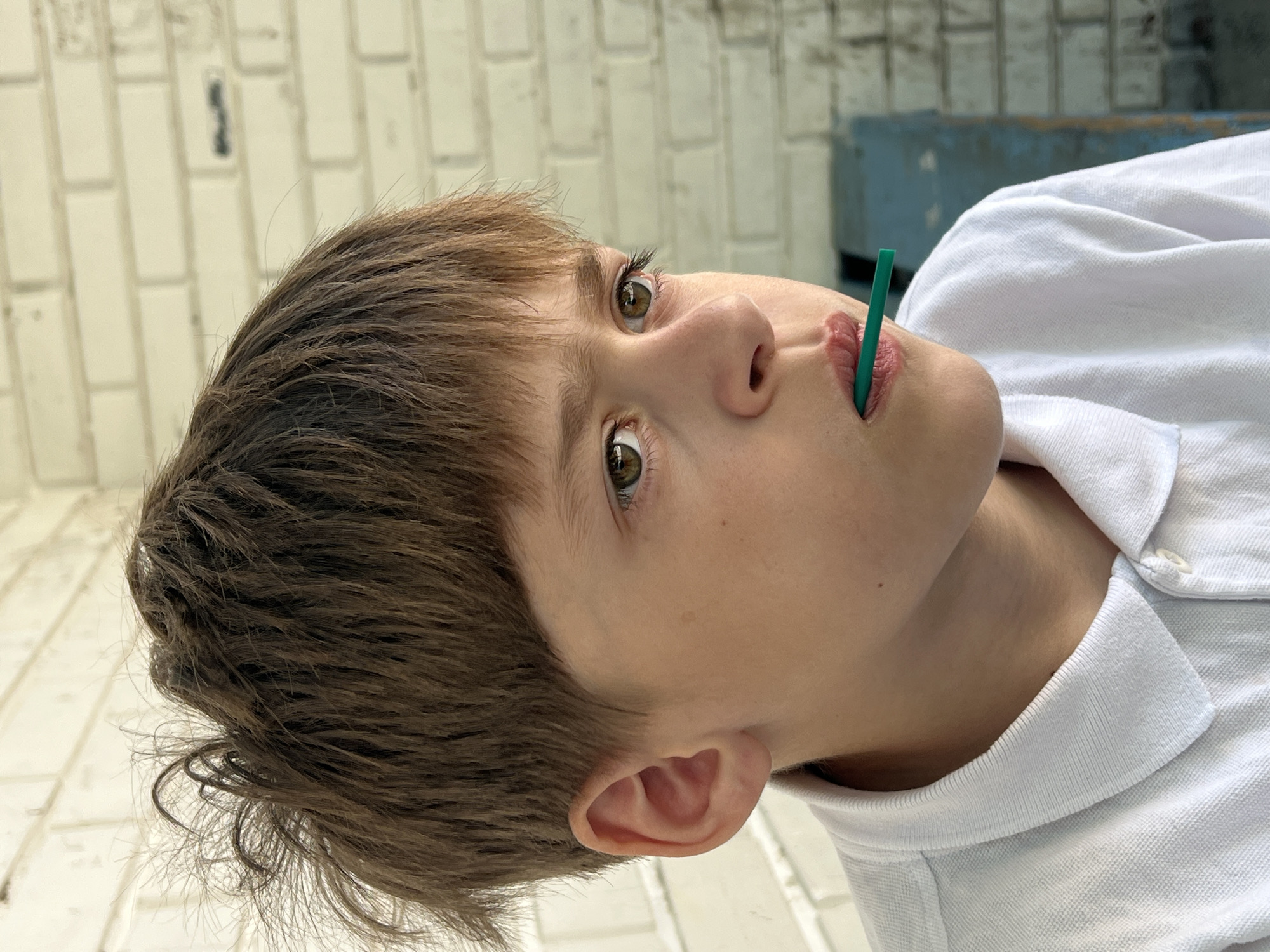 A close-up, tilted portrait shows a young person with fair skin, light brown hair, and hazel eyes. A green straw is positioned in their mouth, with the head turned slightly upwards and to the right. The person is wearing a white collared shirt. The background features a white brick wall and a blue box-like structure partially visible in the upper right. The location of Noviy, Russia is not identifiable in this image.