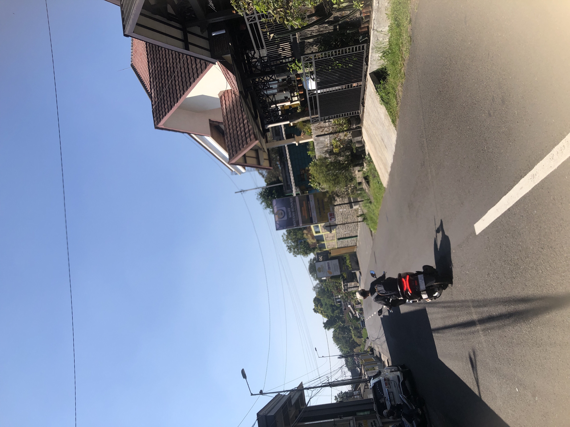 The image depicts a street scene in Trawas, Indonesia, under a clear blue sky. In the foreground, a person is riding a dark-colored scooter on an asphalt road. The rider, wearing a backpack and a light-colored cap, is moving away from the camera. The scooter has a red taillight. To the left of the road, a white SUV is parked, with another motorcycle beside it. Several utility poles with numerous wires stretch across the scene, indicating urban infrastructure.
On the right side of the road, there is a residential building with a tiled roof and white and beige walls. A black metal fence encloses the property, which features potted plants and a concrete walkway. Further down the street, more houses and trees are visible, suggesting a suburban or semi-rural environment. Several signs are placed along the road, one of which appears to be advertising. The overall impression is a sunny day in a developing area, with typical traffic and residential architecture.