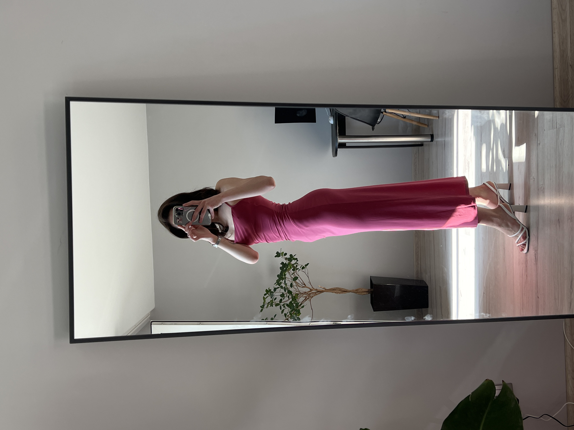 A woman stands in a room, reflected in a full-length mirror, taking a selfie with her phone. She is wearing a long, pink, spaghetti strap dress and white heeled sandals. In the reflection, a tall, black potted plant with thin branches and green leaves stands to her left. Behind the plant, a small, round table with a black top and metal legs is visible, with a black chair positioned nearby. The floor is wooden, and sunlight streams in from the right, creating distinct shadows. The walls are a neutral gray color. The scene appears to be an indoor domestic setting, likely a bedroom or dressing area, possibly in Ternopil, Ukraine.