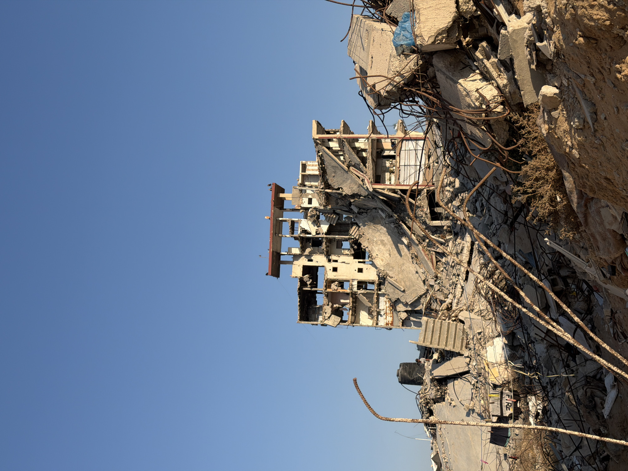 In Jabalia, Palestinian Territories, a multi-story building stands in ruins against a clear blue sky. The structure, once a building, is now a shell of its former self, with floors collapsed and walls exposed, revealing the skeletal framework of rebar and concrete. Debris is scattered across the landscape, a chaotic mix of concrete chunks, twisted metal, and building materials. A rusty lamppost leans precariously to the left, its curved arm reaching towards the sky. In the foreground, piles of rubble dominate, interspersed with sparse, dry vegetation. A black water tank is visible near the base of the ruined building, and a person can be seen standing near it, appearing small against the immense destruction. The scene is stark and desolate, a testament to the destructive forces that have impacted this urban environment.