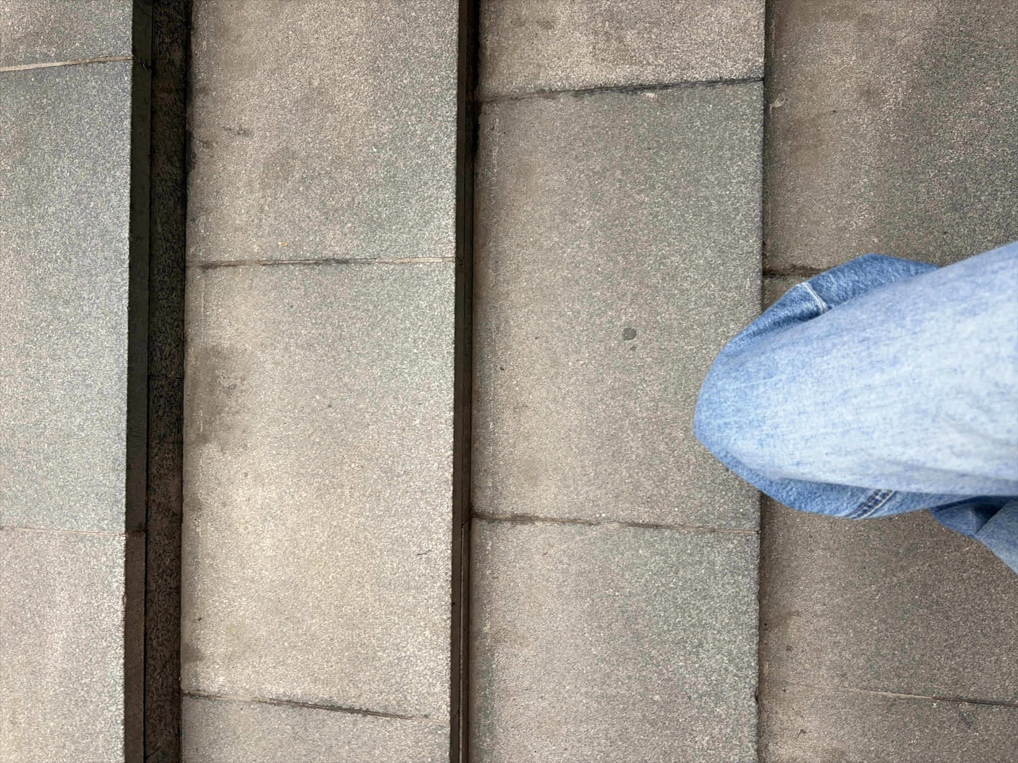 The image shows a top-down view of a person's legs, clad in light blue denim jeans, standing on a set of outdoor steps. The steps are constructed from large, grey stone tiles, with visible grout lines. The tiles have a speckled texture. The person's legs are positioned towards the bottom center of the frame, obscuring the lower portion of the steps. The lighting suggests an overcast day. There are no other visible subjects or objects in the scene. The setting appears to be a public outdoor space, potentially a park or a public building's entrance, within the city of Moscow, Russia.