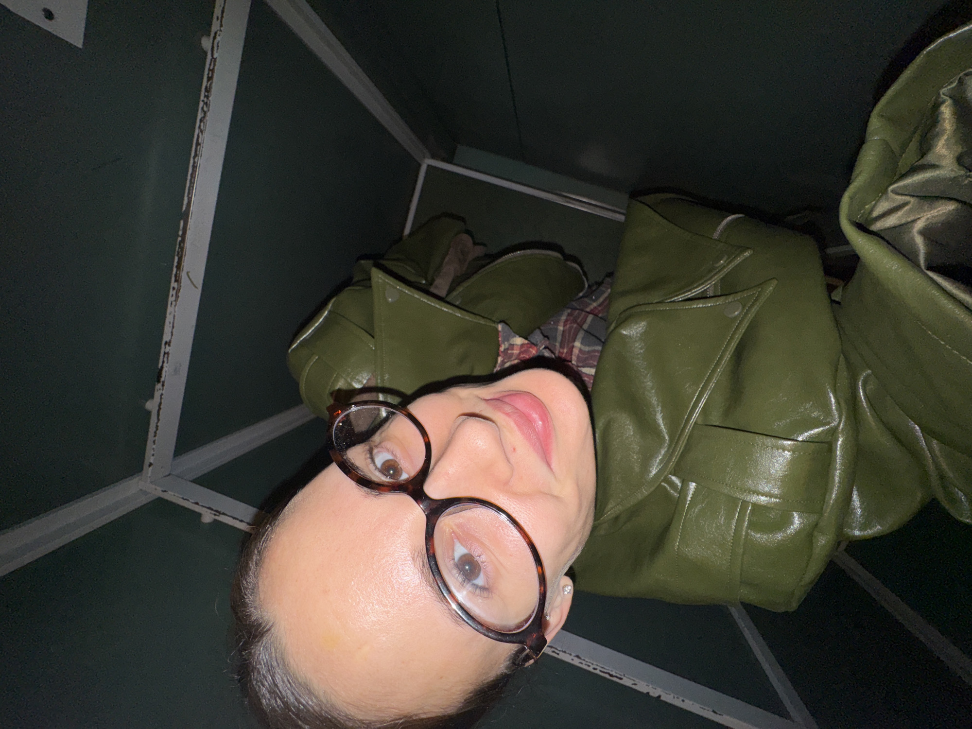 A woman wearing glasses and a green leather jacket is looking up at the camera, which is angled downwards. She is positioned inside a box-like structure with a green interior and white supports, likely a small enclosure or lift in Chaikovsky, Russia. The lighting is dim.