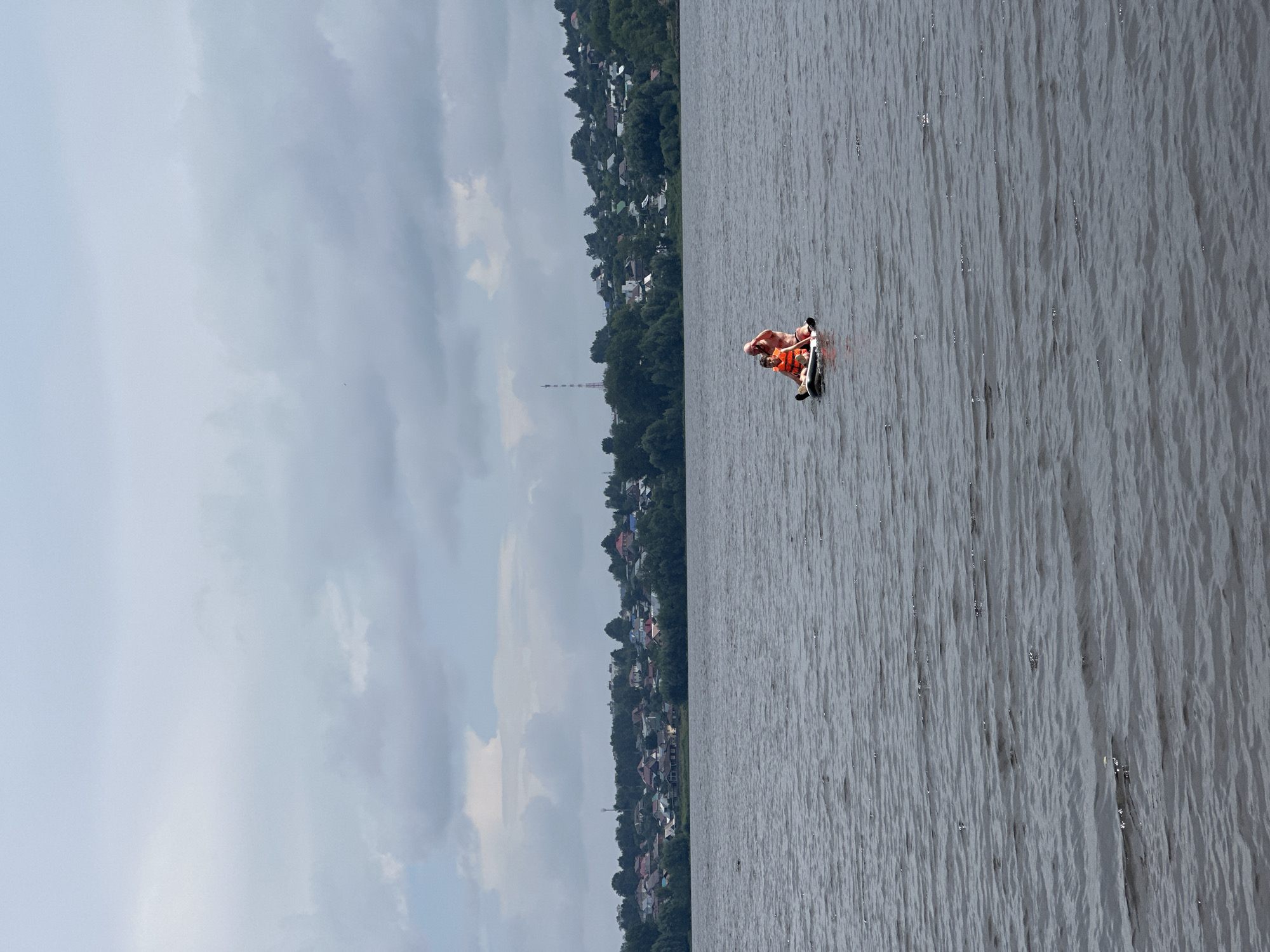 A father and son, both wearing orange life vests, are paddling a small inflatable boat on a wide, choppy river. The father is seated in the back of the boat, and the son is in front. In the background, a treeline of dark green trees separates the river from a hillside covered with a densely packed residential area of houses with red and light-colored roofs. A tall, narrow tower is visible on the hillside. The sky is overcast with thick, gray clouds. The river water is dark gray with small waves, and the overall impression is a cloudy, temperate day. The scene appears to be located on the Kama River in the city of Chaikovsky, Russia.