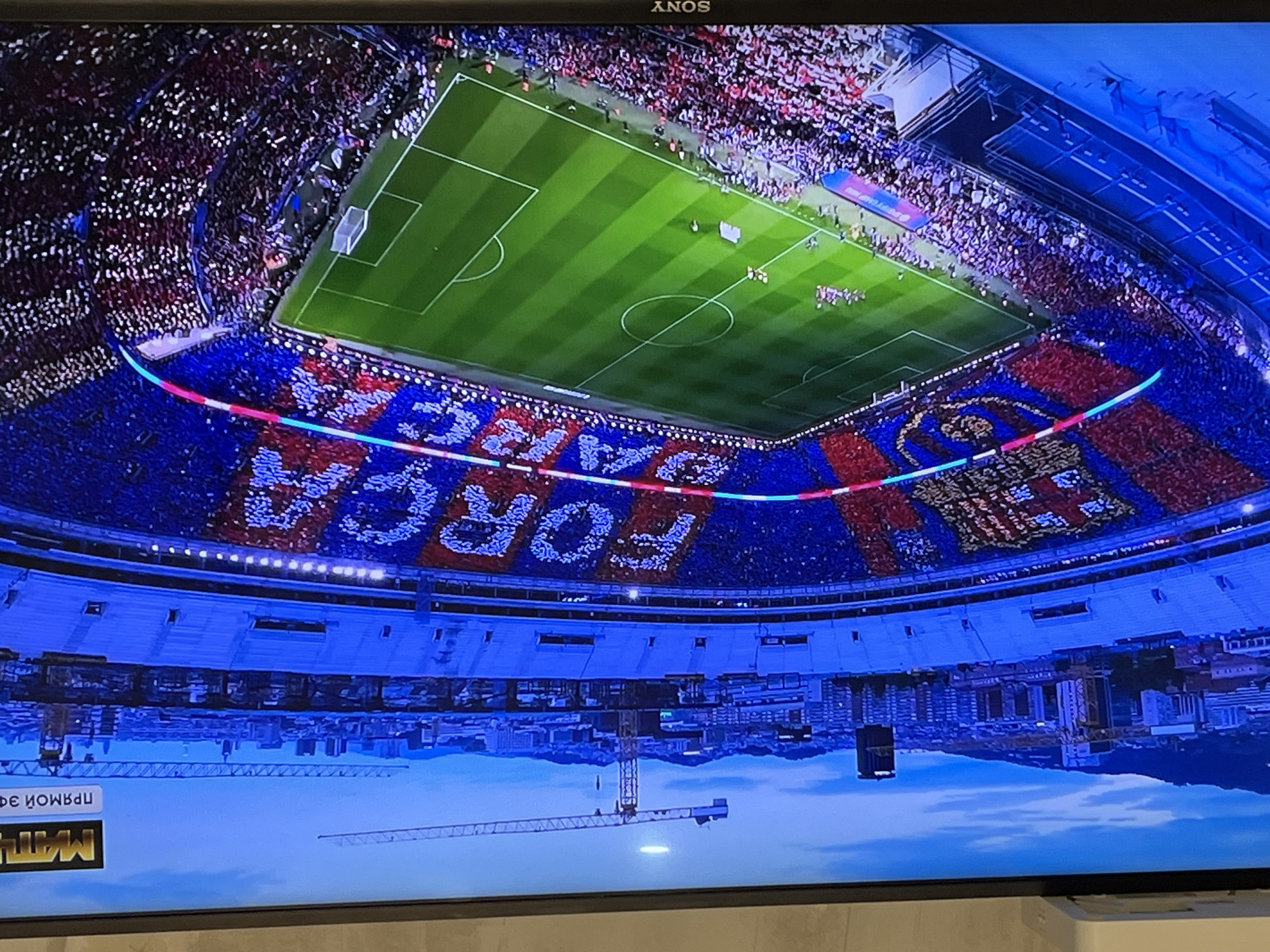 The image shows a television screen displaying an overhead view of a soccer stadium filled with spectators. The stadium appears to be in Barcelona, Spain, evident from the words "Forca Barca" displayed by the crowd and the team's colors. The soccer field is green with white lines. The television's reflection shows a cityscape, including a crane. The television is an Años brand. Text in Russian is partially visible in the lower left corner of the screen.
The scene is not located in Chaikovsky, Russia; it is a broadcast of a soccer match, likely being viewed in an undetermined location.
