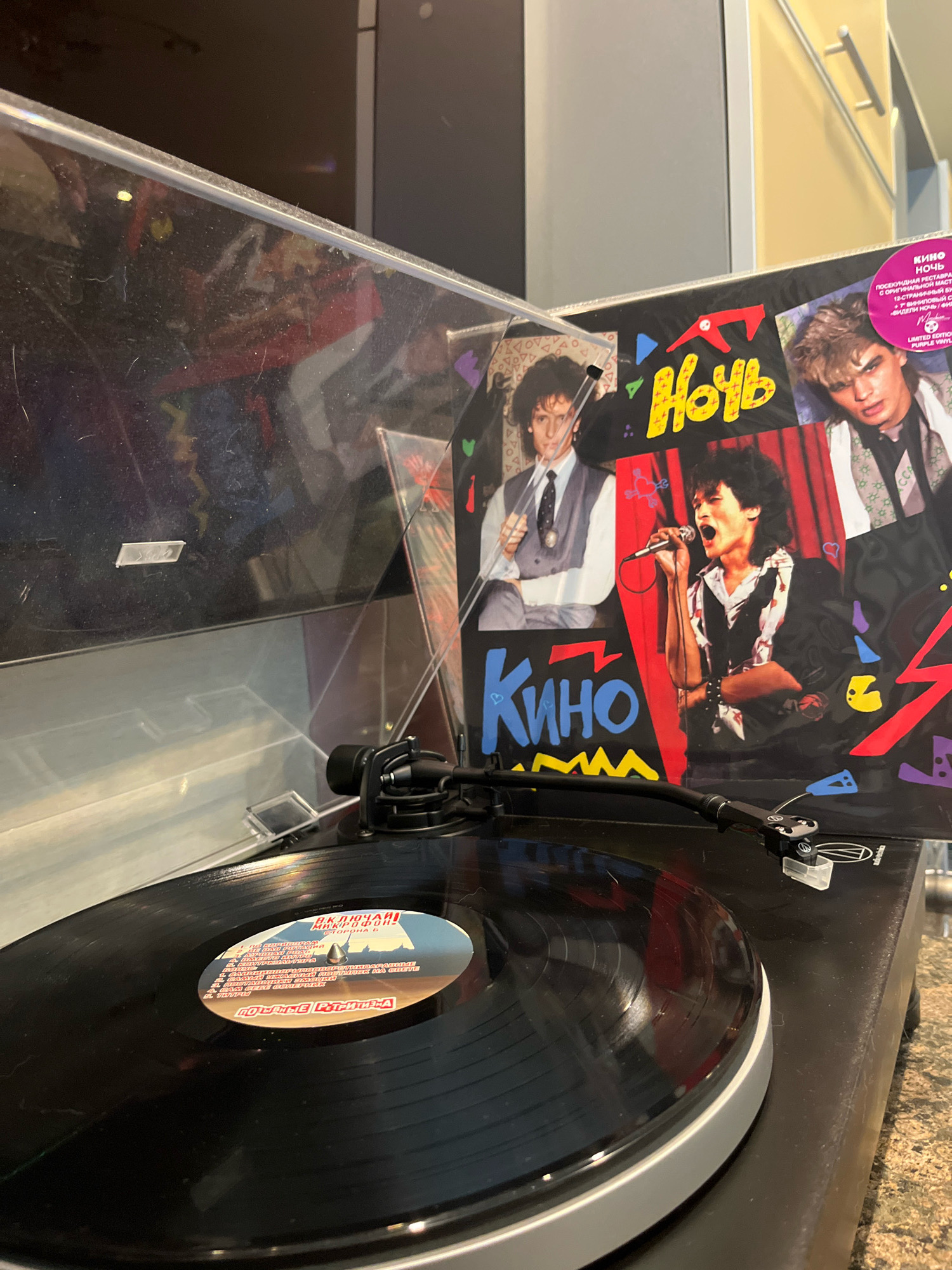 The image depicts a close-up, eye-level shot of a turntable playing a vinyl record. The turntable is positioned on a dark-colored surface, possibly a countertop, with a speckled brown and grey texture. A record sleeve, featuring images of a band and bold text in Russian, leans against the turntable's dust cover. The text on the sleeve is primarily in red and yellow, with the word "Ночь" (Night) prominently displayed. A circular purple sticker is affixed to the upper right corner of the sleeve, also with Russian text. The vinyl record itself is black and partially visible on the turntable platter. The label on the record is circular and features an illustration of a cityscape and text in Russian. The background is blurred, but appears to be an interior setting with hints of furniture and possibly a light fixture. The scene does not provide any specific details that would allow for its location in the city of Chaikovsky, Russia to be identified.