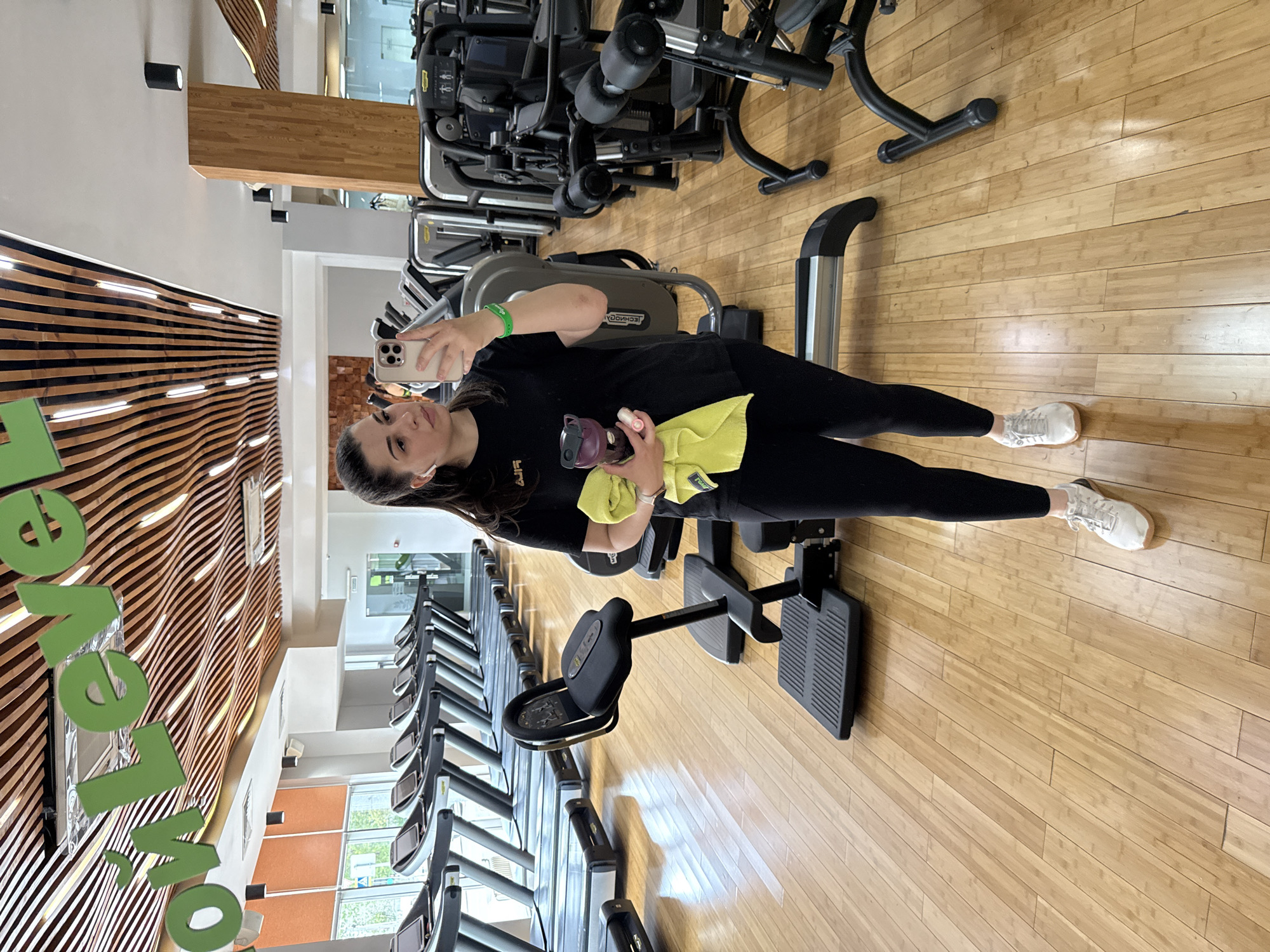 A woman is taking a selfie in a gym. She is wearing black leggings, a black t-shirt, and white sneakers. She is holding a phone in her left hand, and a bottle and a yellow towel in her right hand. She also wears a green bracelet on her left wrist. The gym has several treadmills and other exercise equipment. The floor is made of wood, and the walls are decorated with wooden slats. There are big, light green letters on the wall spelling "OVER." The gym is in Krasnoyarsk, Russia, based on the location tag "Krasnoyarsk" in the image.