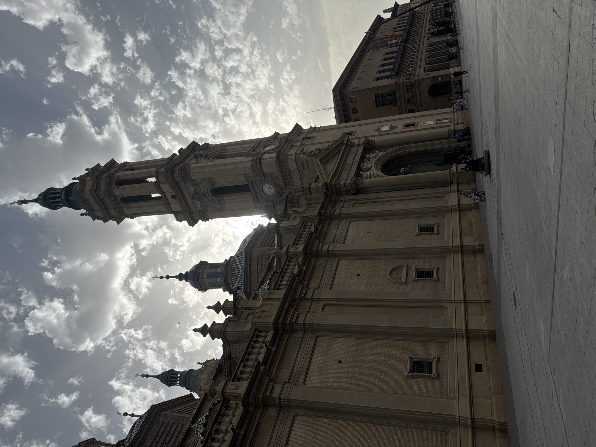 The image shows the Basilica of Our Lady of the Pillar in Zaragoza, Spain. The facade of the basilica is made of brick and stone, with a tall bell tower. The sky is cloudy with the sun shining through. In front of the basilica, there is a large plaza with a few people walking around. The lighting suggests it is either early morning or late afternoon.