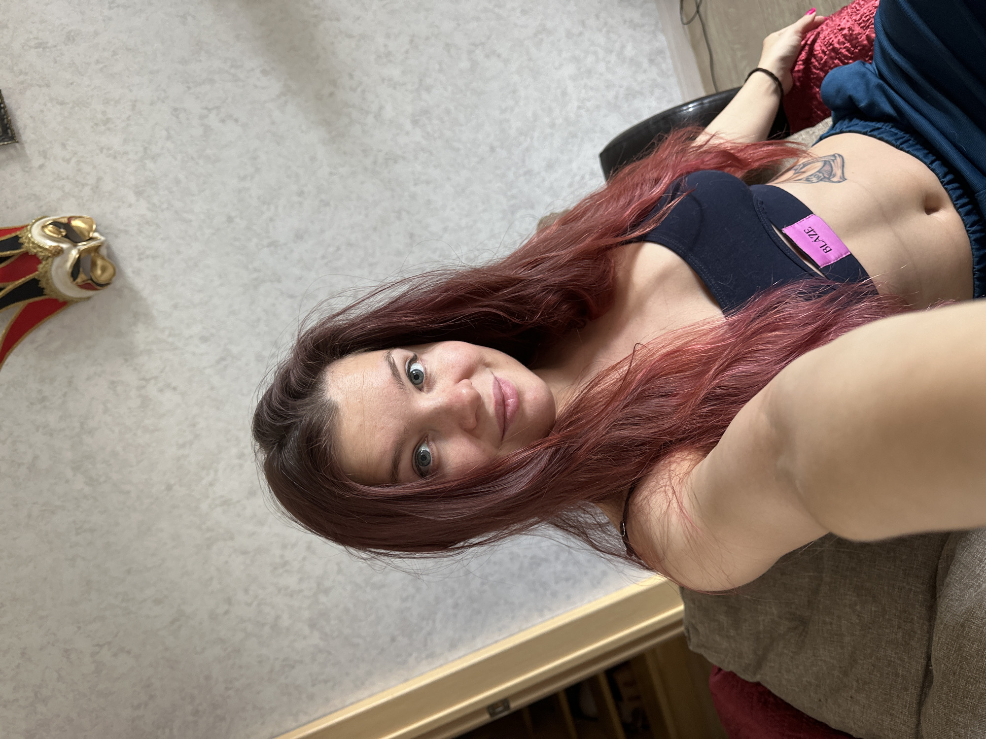 A woman with red hair, wearing a dark bra and blue pants, poses for a selfie. She has a tattoo on her stomach and a sticker reading "BLAZE" on her bra. She is sitting in a chair and the background shows a wall and a cabinet. The location is not identifiable as Vladimir, Russia.