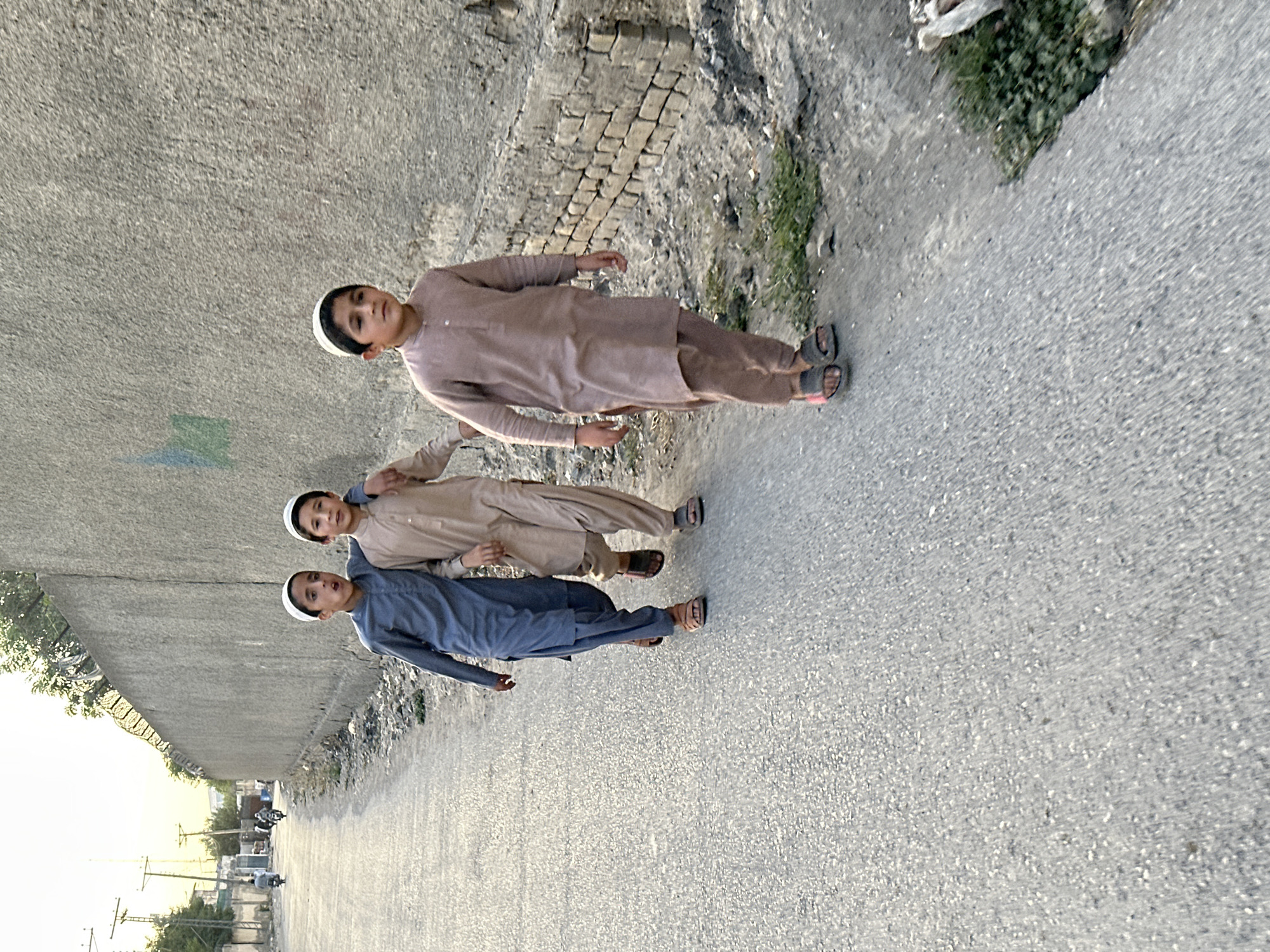 Three boys stand side-by-side on a paved road within Quetta City Tehsil, Pakistan. They are dressed in traditional South Asian clothing and wearing white skullcaps. The boy on the left is wearing a blue outfit, the middle boy is wearing tan, and the boy on the right is wearing a light brown outfit. The road is lined on one side by a wall and vegetation. In the distance, there are buildings, power lines, and vehicles.