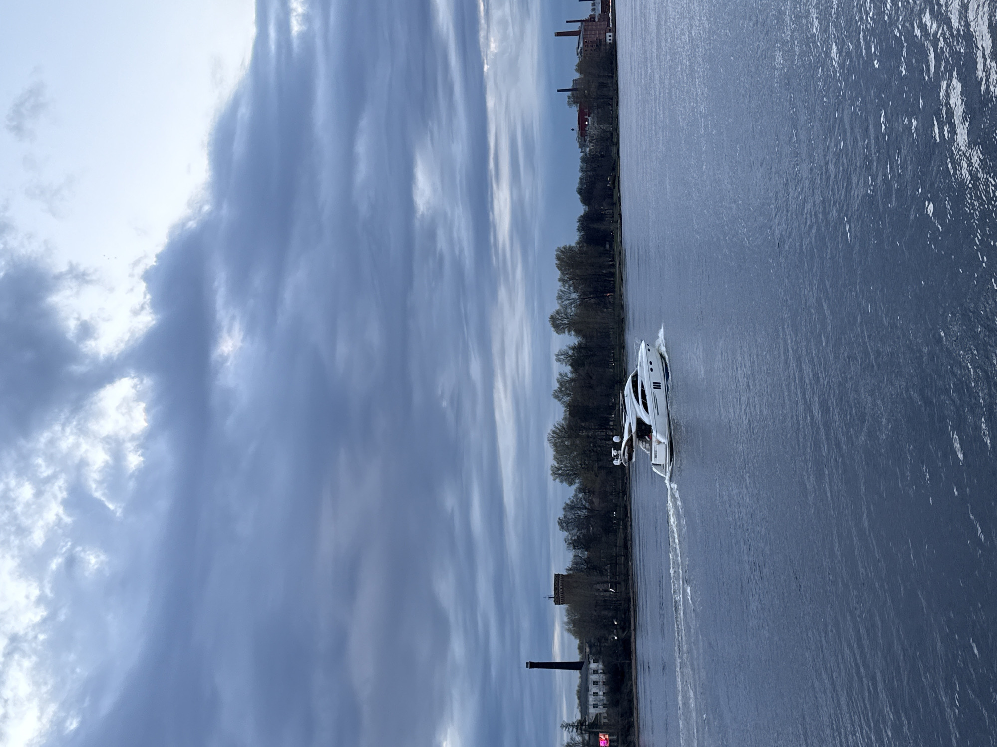 The image displays a wide river or canal in Saint Petersburg, Russia, under a dynamic, partly cloudy sky. The sky is filled with sweeping streaks of clouds, varying from bright white where light breaks through to deeper grey-blue, suggesting either an overcast day or the changing light of late afternoon.
A sleek, white motor yacht or boat is the central subject on the water, moving from right to left across the frame. It leaves a long, foamy white wake trailing behind it, indicating its propulsion. The water itself is a dark, cool blue-grey, showing subtle ripples and reflections of the sky above.
On the distant bank, a dense line of trees, likely deciduous and still largely bare or in early spring, stretches across the horizon, forming a dark silhouette against the lighter sky. Behind the treeline, several buildings are visible, consistent with an urban or industrial waterside environment. These include older red-brick structures with multiple chimneys and a distinctive tall, dark industrial chimney. A square, tower-like building can also be seen further along the bank. No people are clearly discernible, nor is any text visible. The overall mood is calm and somewhat reflective due to the muted colors and cloudy weather.