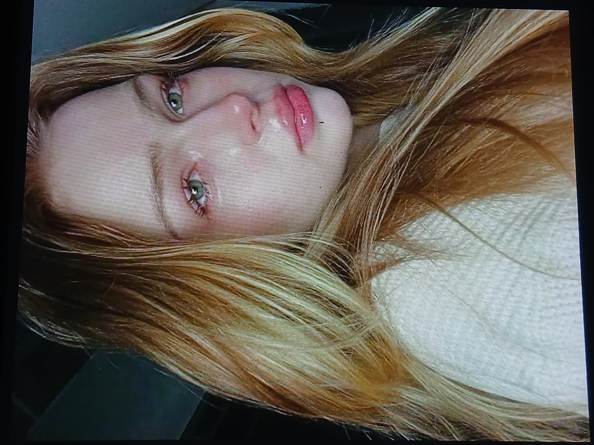 The image shows a close-up, low-angle, slightly blurry, and grainy photograph of a young Caucasian woman with long, wavy, blonde and light brown hair. Her eyes are green and appear to be red-rimmed and watery, with visible tears streaming down her face and glistening on her cheeks. She has full, pink lips, which also appear glossy. She is wearing a light-colored, textured sweater. The background is dark and indistinct, with some shapes that might suggest an indoor environment, possibly a car interior given the dark, angled shapes. The lighting is dim, with a focus on the woman's face. There are no discernible landmarks or contextual clues to indicate a specific location within the city of Dnipro, Ukraine.