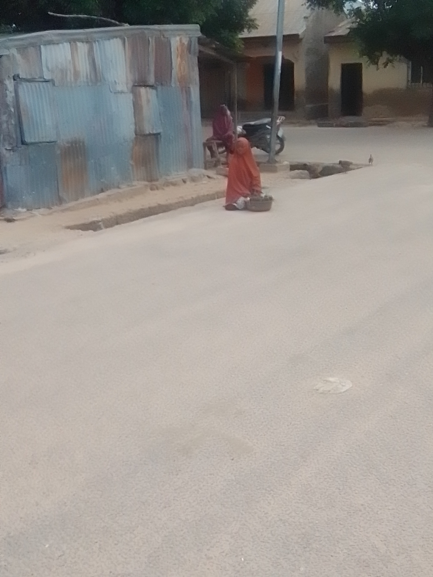 In Liji Hill, Nigeria, a woman in an orange garment is crouched on the side of a dirt road, tending to a basket at her feet. Behind her, a motorbike is parked near a building with two dark doorways. To the left, a corrugated metal structure stands against a backdrop of trees and other buildings. A single bird is visible in the distance on the road. The ground appears to be dusty, with a few white splotches on the road surface.
