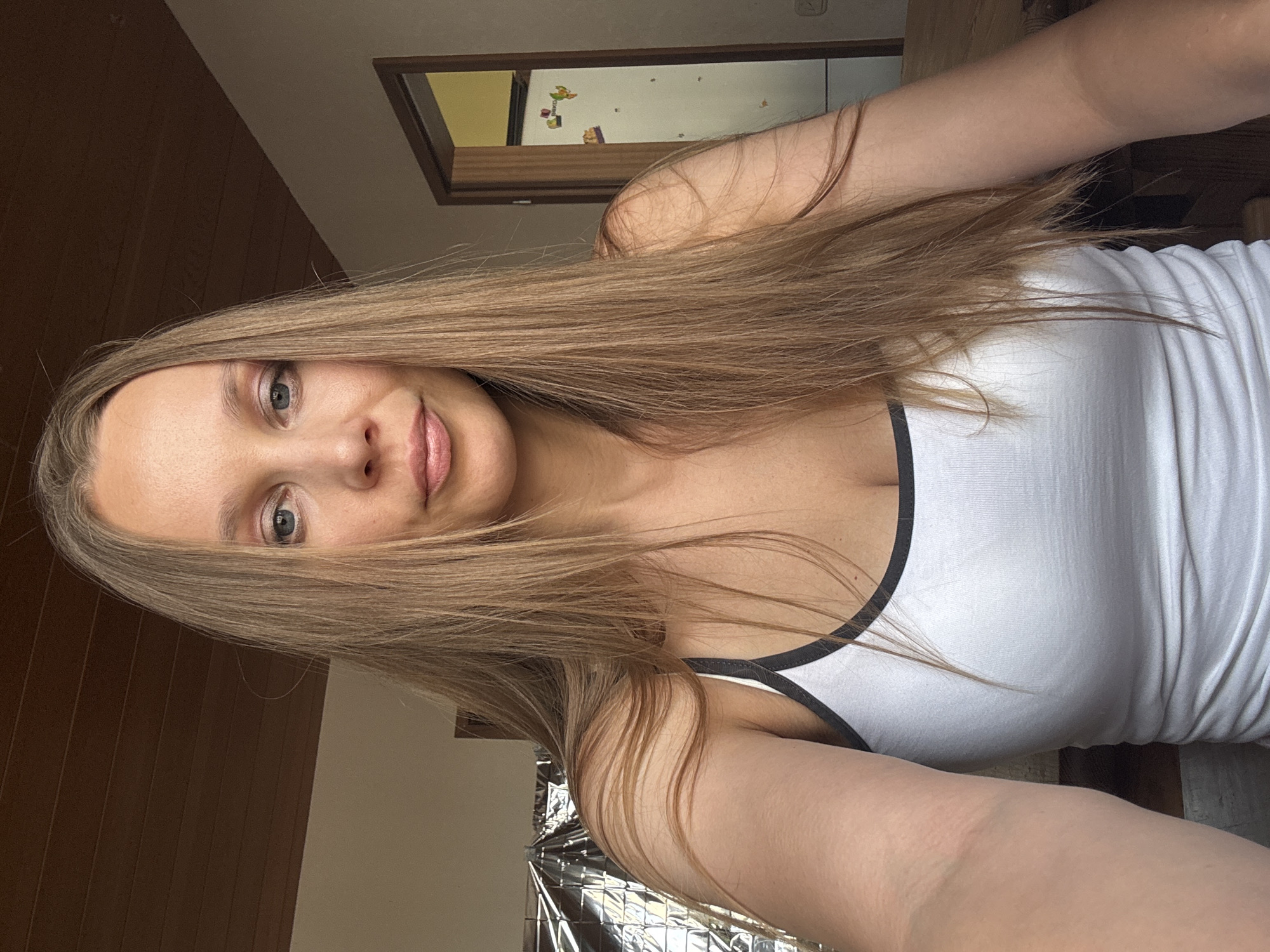 A young woman with long, straight blonde hair is pictured indoors, taking a selfie. She is wearing a white tank top with black trim. Her face is turned slightly towards the camera, and she is looking directly at it. She has blue eyes and light makeup. The background shows a ceiling with wooden planks, a wall, and a doorway to another room. The room appears to be a residential interior, possibly a home. There are no immediate indicators of the specific location within Überherrn, Germany, beyond it being an interior space.