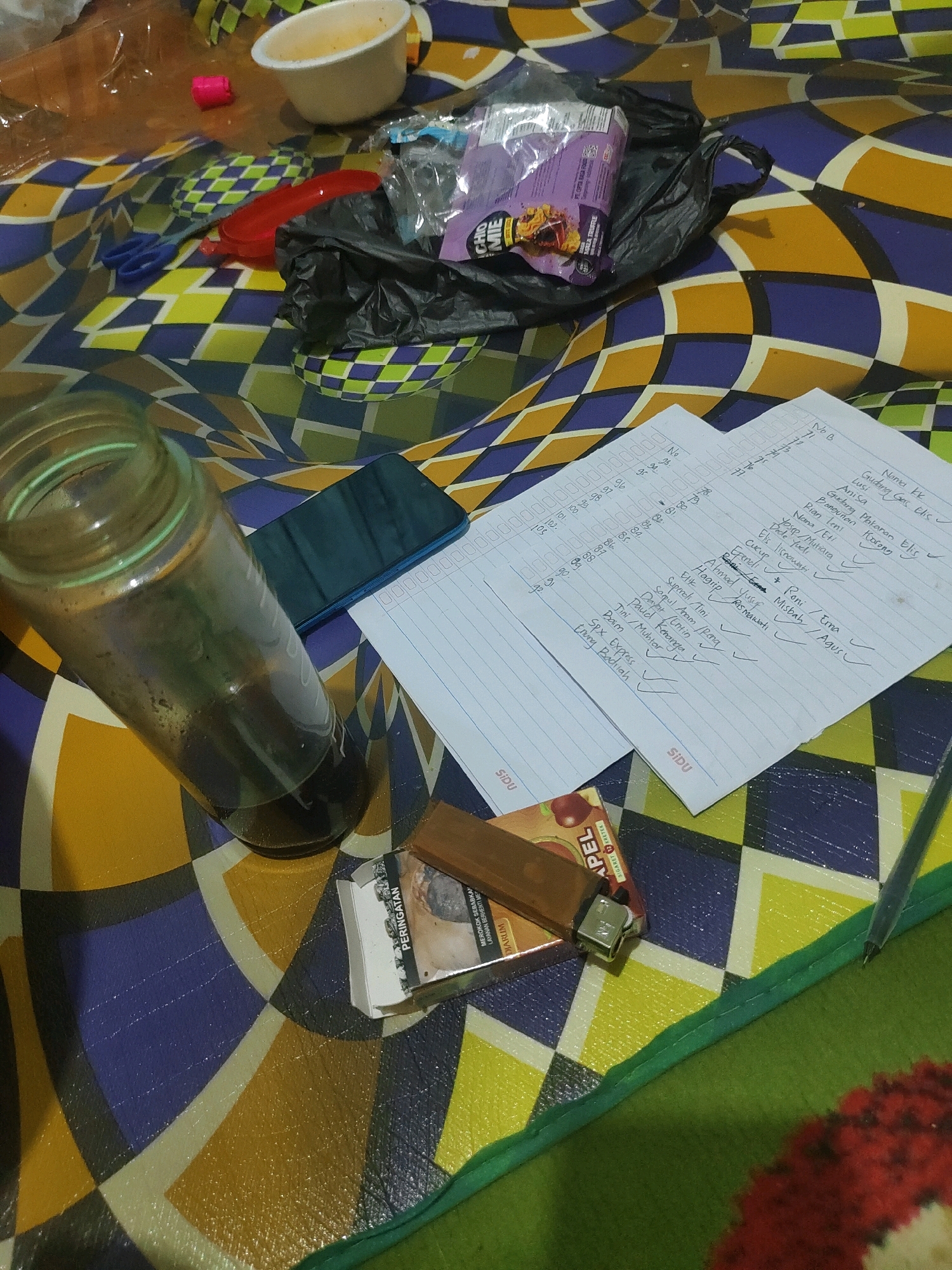 The image depicts a close-up, overhead view of a cluttered surface, likely a floor or table, adorned with a colorful, geometric patterned fabric. The objects on the surface suggest a casual, perhaps late-night, study or work session.
Prominently featured is a partially filled glass bottle containing a dark liquid, possibly coffee or tea. Next to it, a blue smartphone rests on its side. Two sheets of white paper, covered in handwritten notes and lists, are laid out, with one sheet containing a list of names and checkboxes.
Other items include a pack of cigarettes, a lighter, a small red plastic object, a white bowl, a pair of blue scissors, and a black plastic bag containing what appears to be a food item in purple packaging. A red plastic scoop is also visible.
The overall impression is one of disarray, with personal effects scattered around. There are no people visible in the image. The environment appears to be indoors, with the lighting suggesting it might be artificial. The mention of Sukanagara, Indonesia, suggests a possible geographical context for this scene.
