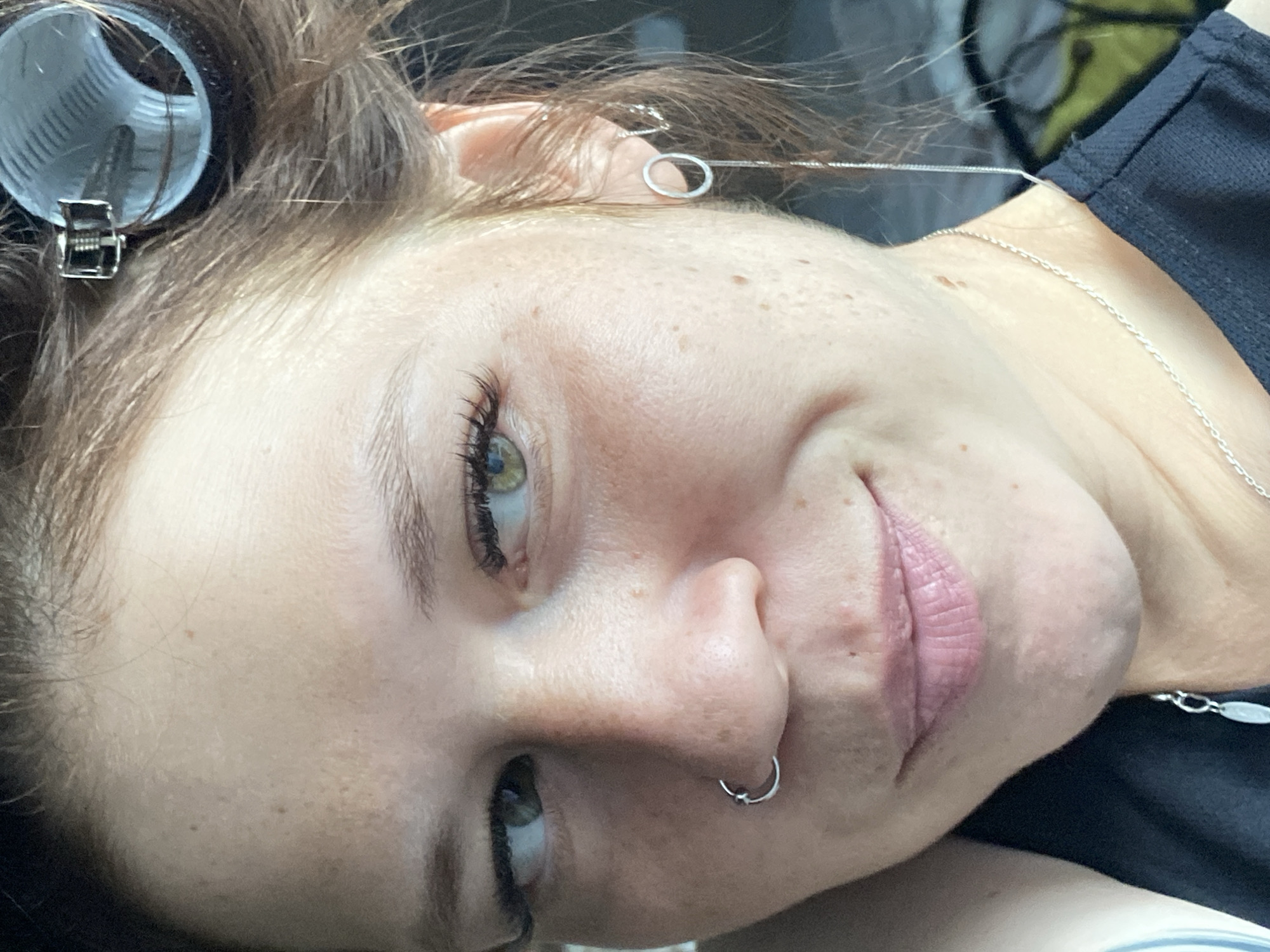 A close-up, low-angle medium shot shows a woman with light skin and green eyes. She has freckles scattered across her face and nose. Her left eye is looking slightly to the left, while her right eye is looking straight ahead. Her lips are closed and appear to be wearing a light pink lipstick. She has a silver hoop piercing in her left nostril.
Her hair is brown and styled with a large, blue hair roller visible on the right side of her head, secured with a silver clip. She is wearing a black top with a delicate silver necklace around her neck, which has a pendant that is partially visible. On her right ear, she wears a silver earring that consists of a small hoop with a long, delicate chain dangling from it. The background is blurred and appears to be indoors, with hints of fabric and possibly furniture. The lighting suggests it is daytime, with soft light illuminating the woman's face from the front and slightly to the side. The overall scene is intimate and personal, capturing a moment of personal grooming or preparation.