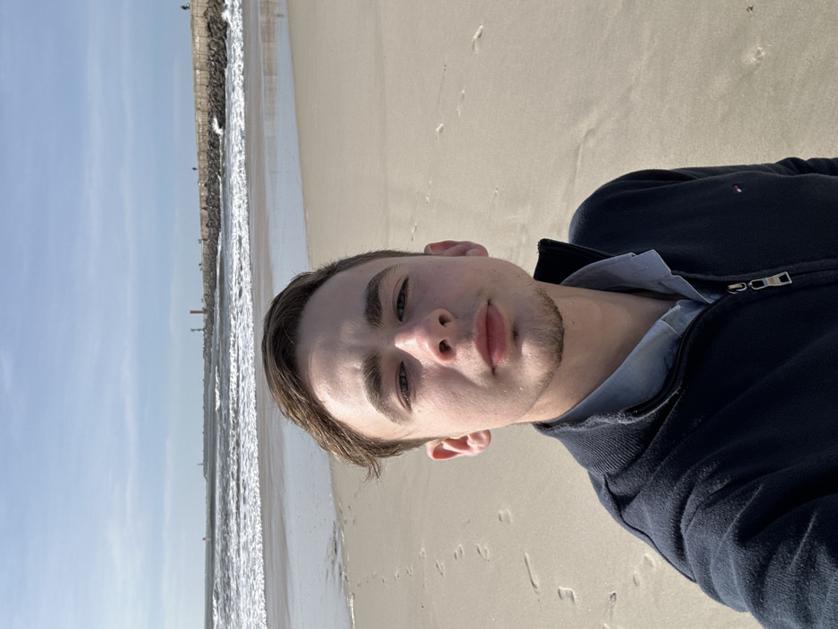 A young male with light skin, short brown hair, and stubble is depicted in a selfie, centrally positioned and looking directly into the camera. He wears a dark blue zippered fleece jacket over a light blue collared shirt. The setting is a sandy beach in Gafanha da Nazaré, Portugal. In the background, ocean waves break on the shore adjacent to a long, grey stone pier extending into the water. Two lighthouses, one red and one white, are visible along the pier's length, with several indistinct human figures also present on the pier in the distance. The sandy foreground displays scattered footprints. The sky is partly cloudy with patches of blue.