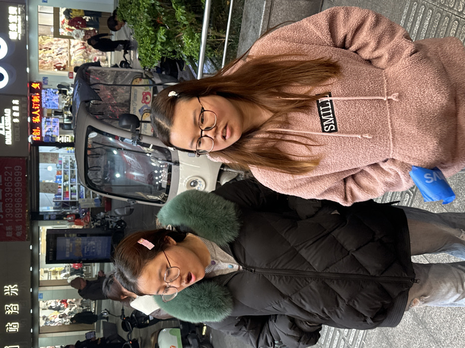 The image depicts two young East Asian women standing on a paved sidewalk in an urban area of Jiangjin, China, during what appears to be evening or night, illuminated by artificial light.
The woman on the left wears a black quilted puffer jacket with a prominent teal-colored faux fur collar over a light grey zipped sweater and light-colored jeans. She has dark hair, wears round, light-framed glasses, and a pink rectangular hair clip on the right side of her head. Her mouth is open as if speaking or reacting.
The woman on the right wears a pink textured hooded sweatshirt with a central front pocket and a black rectangular patch bearing the white word "SMILE" on the chest. She has light brown hair, wears round, dark-framed glasses, and two small white flower-shaped hair clips on the right side of her head. Her mouth is slightly open, and she holds a crumpled blue plastic object in her left hand, which rests near her front pocket. She is looking towards her right.
In the mid-ground, behind the women, a light-colored, three-wheeled, enclosed electric scooter with clear plastic panels and a dark roof is parked. The background features several brightly lit storefronts with Chinese characters, English words such as "DUOLASIGAO" and "SMILE," and displayed phone numbers including "13983396521" and "18996399699." Other indistinct pedestrian figures are visible further back. To the far right, a silver metal railing and green foliage are present.