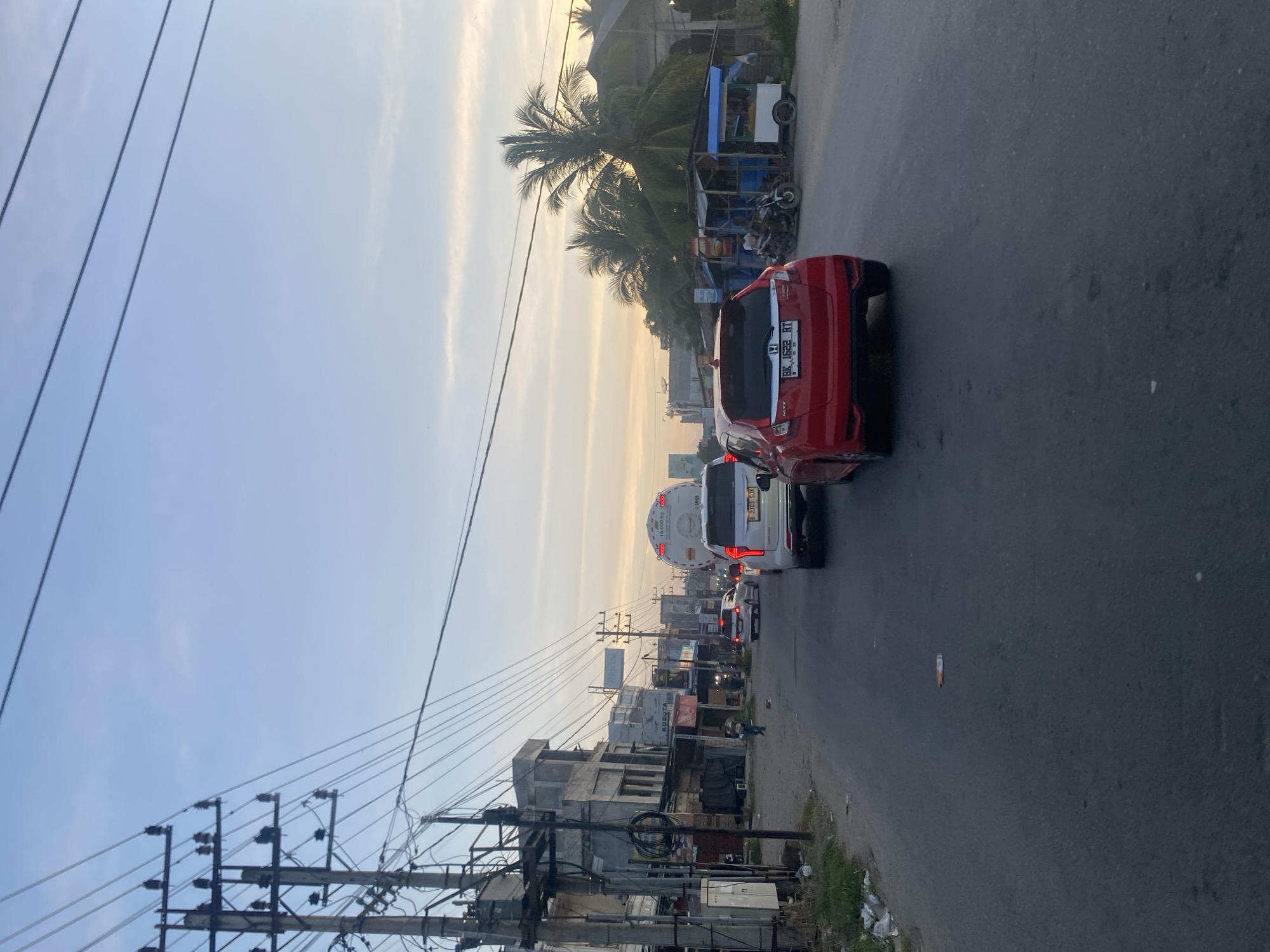 The image depicts a street scene in Tingkeummanyang, Indonesia. Cars, including a red one in the foreground, are traveling on a paved road. Utility poles line the street with many electrical wires. Buildings of various heights and materials are present on the roadside. A palm tree is visible, indicating a tropical climate. The sky is light blue, suggesting daytime. The overall impression is a typical Indonesian urban environment with mixed infrastructure.