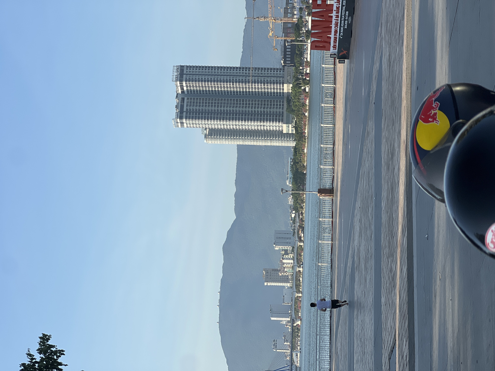 A full shot captures a city skyline during the daytime. In the foreground, two motorcycle helmets, one black and one dark with a red bull graphic, rest on a paved surface. A lone person jogs on a promenade, facing away from the camera and towards a body of water. The water is calm and reflects the sky. A white metal fence borders the promenade. In the middle ground, a city rises with several tall buildings and a bridge spanning the water. In the background, a large mountain range dominates the horizon under a clear blue sky. A large red sign spelling "DANANG" is visible on the right side of the promenade, with smaller text below it. Construction cranes are also visible behind the buildings.