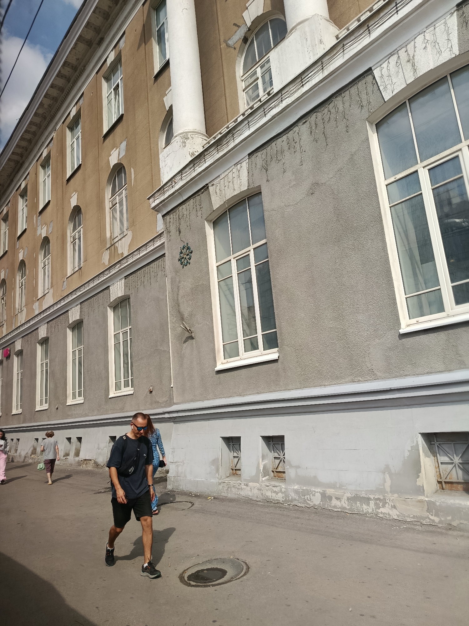 This image captures a street scene in Saratov, Russia. The main subject is a man walking towards the viewer, wearing a dark t-shirt, black shorts, sunglasses, and black athletic shoes. He has a black fanny pack slung across his chest and white earbuds in his ears. Behind him, a woman in a blue patterned dress is also walking away from the viewer. Further in the background, another person in pink is walking away from the viewer, and an older woman with a green bag is walking towards the viewer.
The setting is an urban environment with a large, multi-story building on the left side of the frame. The building has a light brown upper section and a gray lower section, with numerous windows. The architecture suggests a somewhat older, possibly institutional building, with ornate trim and columns. Decorative string lights hang from a ledge along the gray section of the building, and a snowflake-shaped ornament is visible on the wall. The sidewalk or street is paved and shows signs of wear, with cracks and a circular manhole cover in the foreground. The sky above is blue with some clouds, suggesting daytime, likely late morning or early afternoon given the light and shadows. The overall mood is that of everyday urban life, with people going about their business on a sunny day.