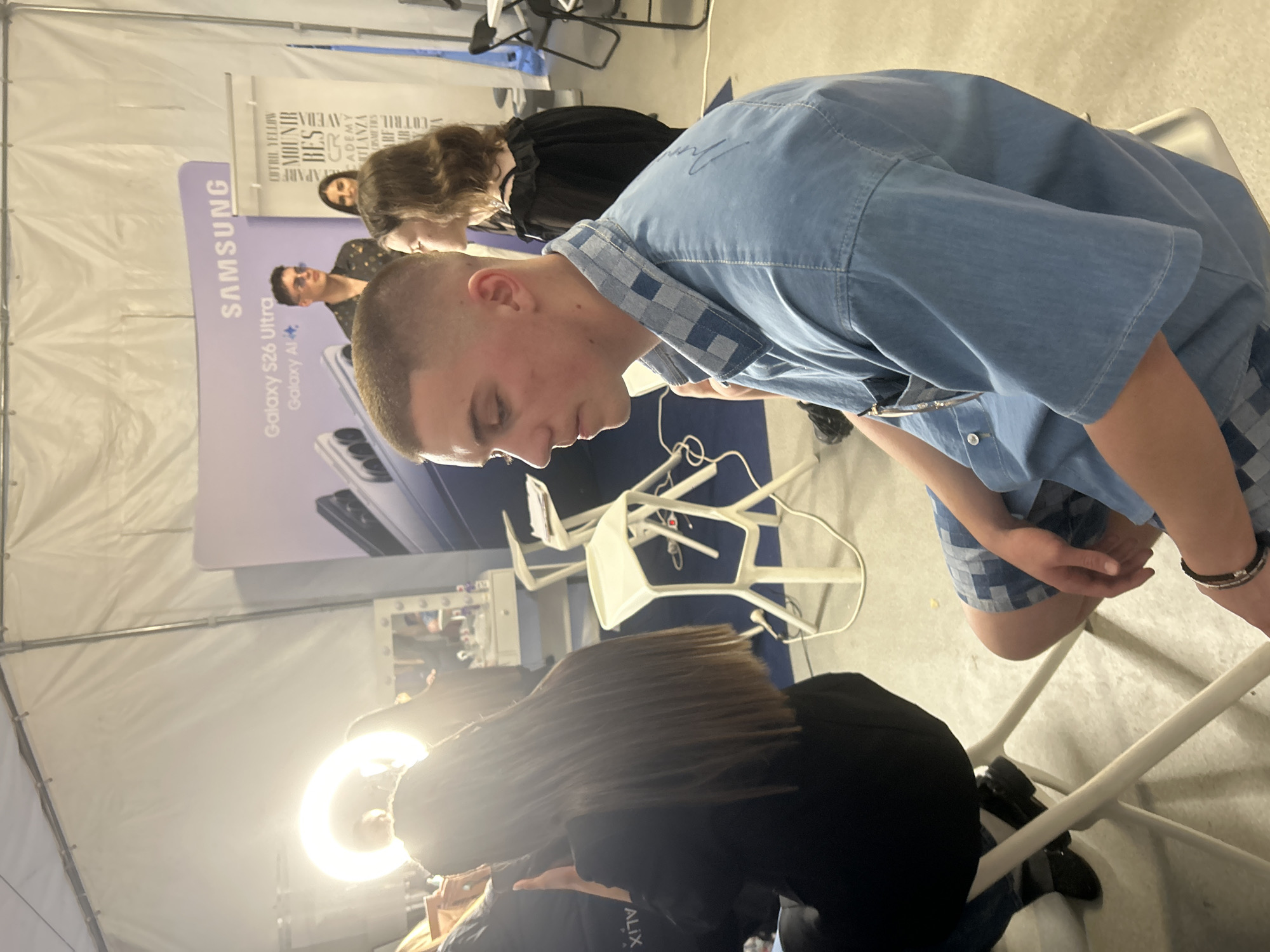 The image depicts an indoor event space, likely a temporary structure such as a large tent, characterized by white canvas walls and ceiling supported by metal framework. The scene is located in the city of Sofia, Bulgaria.
In the foreground, a young male subject with a short, faded haircut is seated in profile, facing right. He wears a blue denim short-sleeved shirt with a checkered collar and matching shorts, and a dark bracelet on his left wrist. To his left, another subject with long brown hair is seen from behind, seated in front of a brightly illuminated ring light, indicative of a grooming or photography session. This subject wears a black top or jacket. Further into the midground on the right, a young female subject is partially visible, looking left, dressed in a black ruffled top. In the background, a male subject wearing sunglasses and a patterned dark shirt stands, observing the scene, with part of another female subject's face visible behind him.
The environment includes a white vanity table with an illuminated mirror and various cosmetic products, flanked by two modern white bar stools. Two prominent branded banners are visible: a purple one for "SAMSUNG Galaxy S26 Ultra" and "Galaxy AI," and a white vertical banner listing several cosmetic and hair brands including "MOUNIR," "BES," "CR ACADEMY," "ATLANZA," "COTRIL," and "WELLA," along with "OUTFIT YELLOW." The floor is light-colored, and various white chairs are distributed throughout the space. The overall arrangement suggests a professional preparation area or promotional media setup for an event.