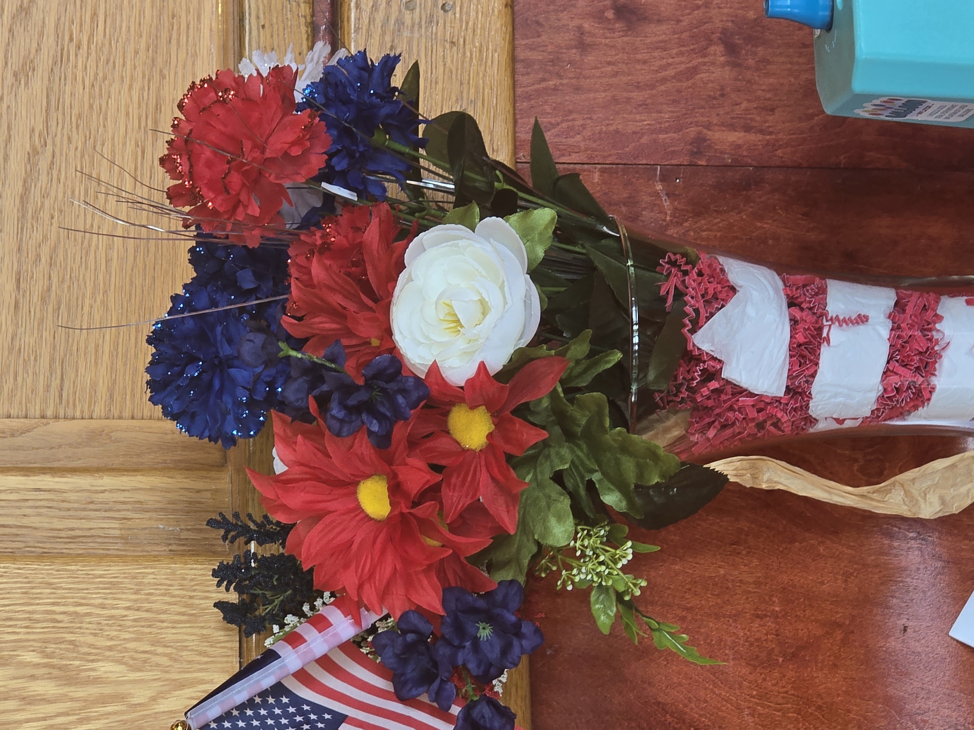 A floral arrangement with American flags is displayed in a glass vase decorated with red and white stripes, sitting on a brown surface against a wooden background. The arrangement features red and blue artificial flowers, including daisies and carnations, with a white rose in the center. A small American flag on a pole is positioned to the left of the flowers. The vase is filled with red shredded paper. The background consists of two wooden panels, one lighter and one darker, suggesting a room interior. The lower right corner shows a portion of a blue object, possibly a dispenser or container. The scene appears to be set up for a patriotic celebration or event. The image was captured indoors, likely in Cumming, Georgia, United States of America.
