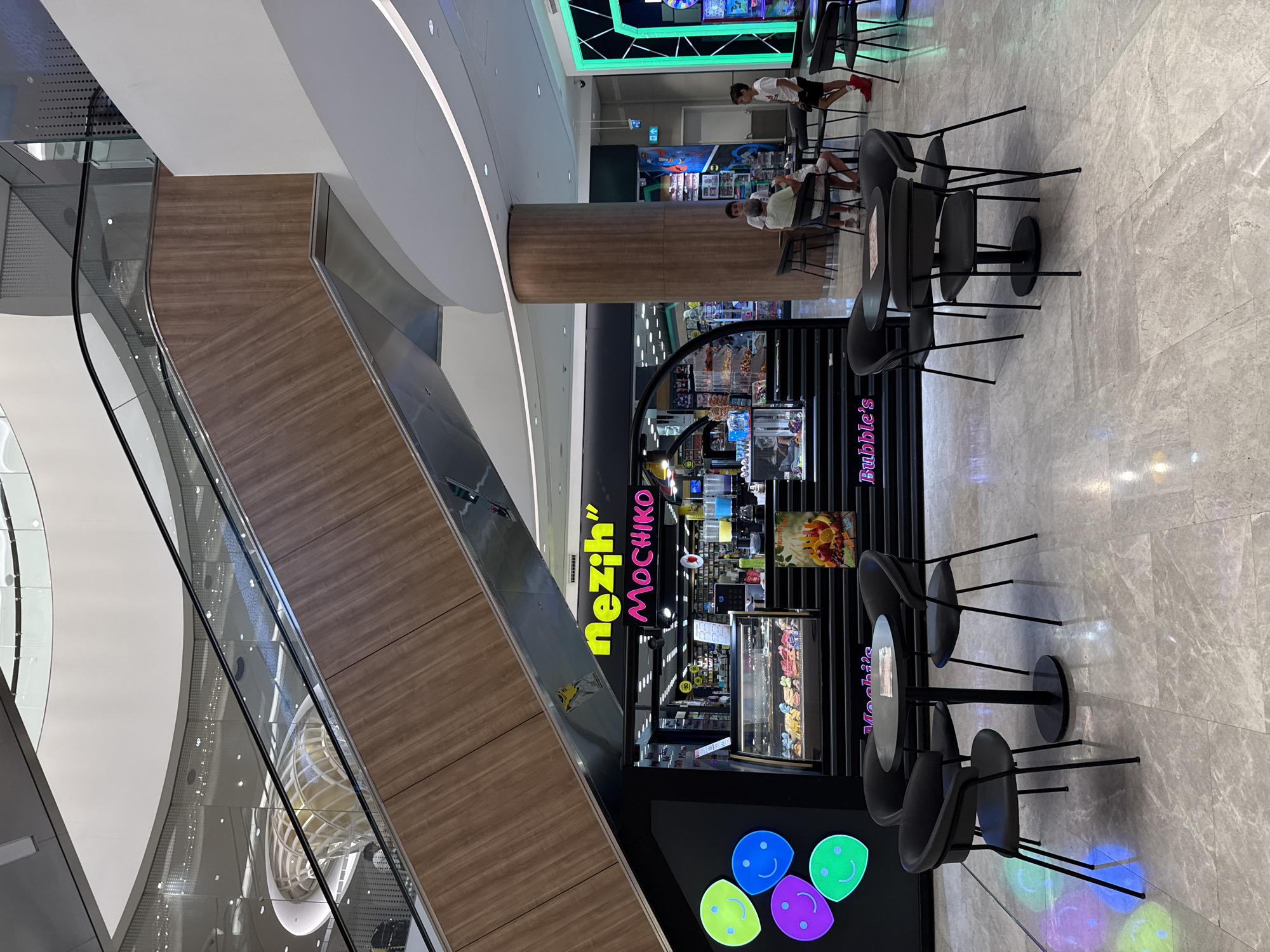 This is a photograph taken inside a modern shopping mall. In the foreground, there are black tables and chairs arranged on a marble-tiled floor. To the left, a storefront named "nezih" and "Mochiko" is visible, featuring a display case filled with colorful pastries and drinks. Above the storefront, a wooden-paneled escalator ascends. In the mid-ground, several people are seated at tables, including a family group. Further back, other shops and escalators are visible within the mall's multi-level interior. The mall features contemporary architecture with white walls, curved ceilings, and ambient lighting. The overall scene depicts a public commercial space. This location is not in Üsküdar, Turkey.