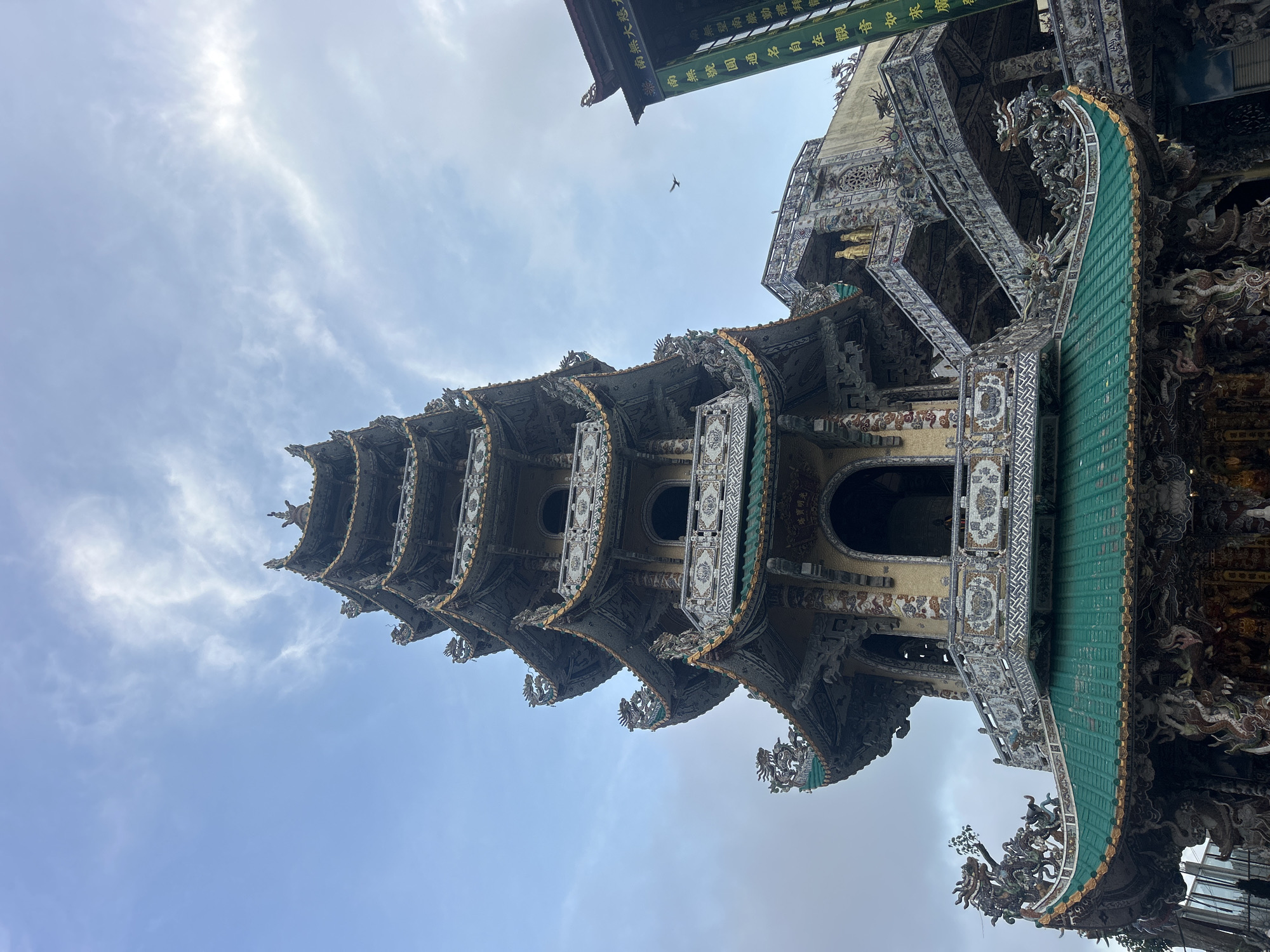 The image presents a low-angle view looking upwards at a multi-tiered, elaborately decorated pagoda or temple building, likely located in Xuân Thành, Vietnam. The structure is characterized by its intricate architecture, featuring multiple levels of ornate roofs, balconies, and walls. The roofs are predominantly green, while the building's facade is adorned with detailed patterns, carvings, and mosaic-like decorations. Numerous mythical creatures, particularly dragons, are visible along the eaves and corners, adding to the grandeur of the design.
The pagoda stands against a bright, partially cloudy sky, indicating daylight. The lighting is soft and even, suggesting either an overcast day or the sun positioned high above. A small bird is captured in flight high in the sky. A green banner or sign with traditional Chinese characters hangs from an upper section of the building. The legible text includes phrases such as "南無大慈大" (Namo Great Compassionate Great), "無望南巖勤禮拜" (No hope, Southern Rock diligently worship), and "南無號圓通名自在觀音如來廣" (Namo Perfectly Penetrating, Self-Existent Avalokiteshvara Buddha, Vast), indicating a Buddhist context and devotion to Guanyin. The upward perspective emphasizes the height and detailed craftsmanship of the religious site. No individuals are visible, allowing the focus to remain on the architectural beauty and the expansive sky.