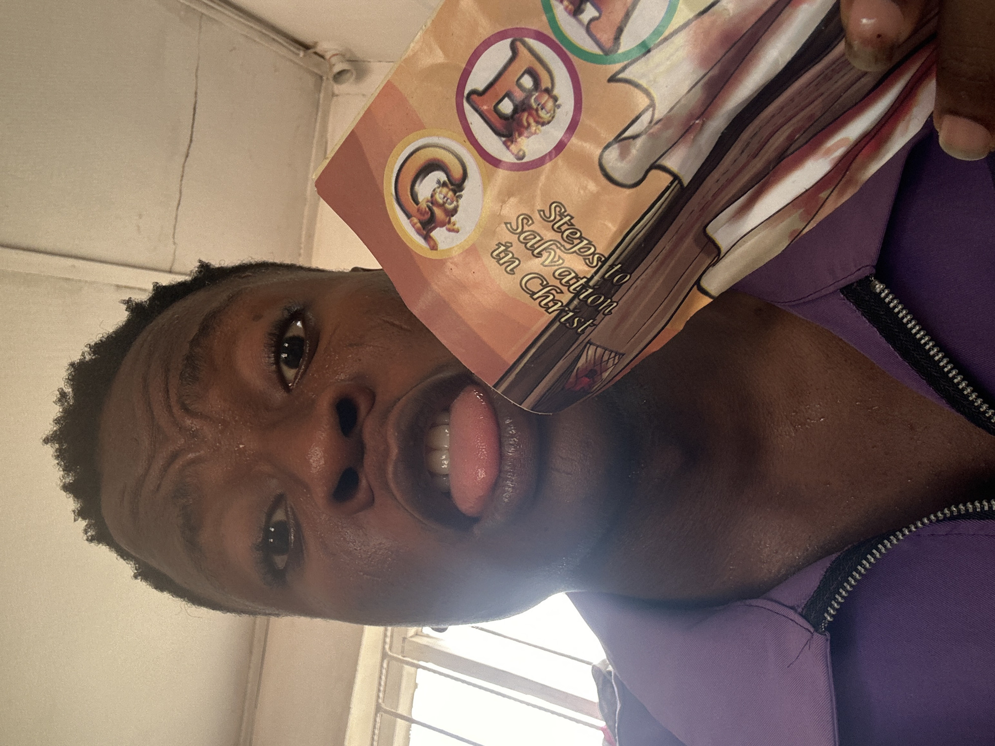A person, presumably male, holds a book titled "Steps to Salvation in Christ" in front of their face. The person is wearing a purple jacket with a black zipper. The background appears to be an interior space with a window. The location of Ikorodu, Nigeria, is not verifiable from the image itself. There are stickers on the book with cartoon animals. The person's tongue is sticking out.