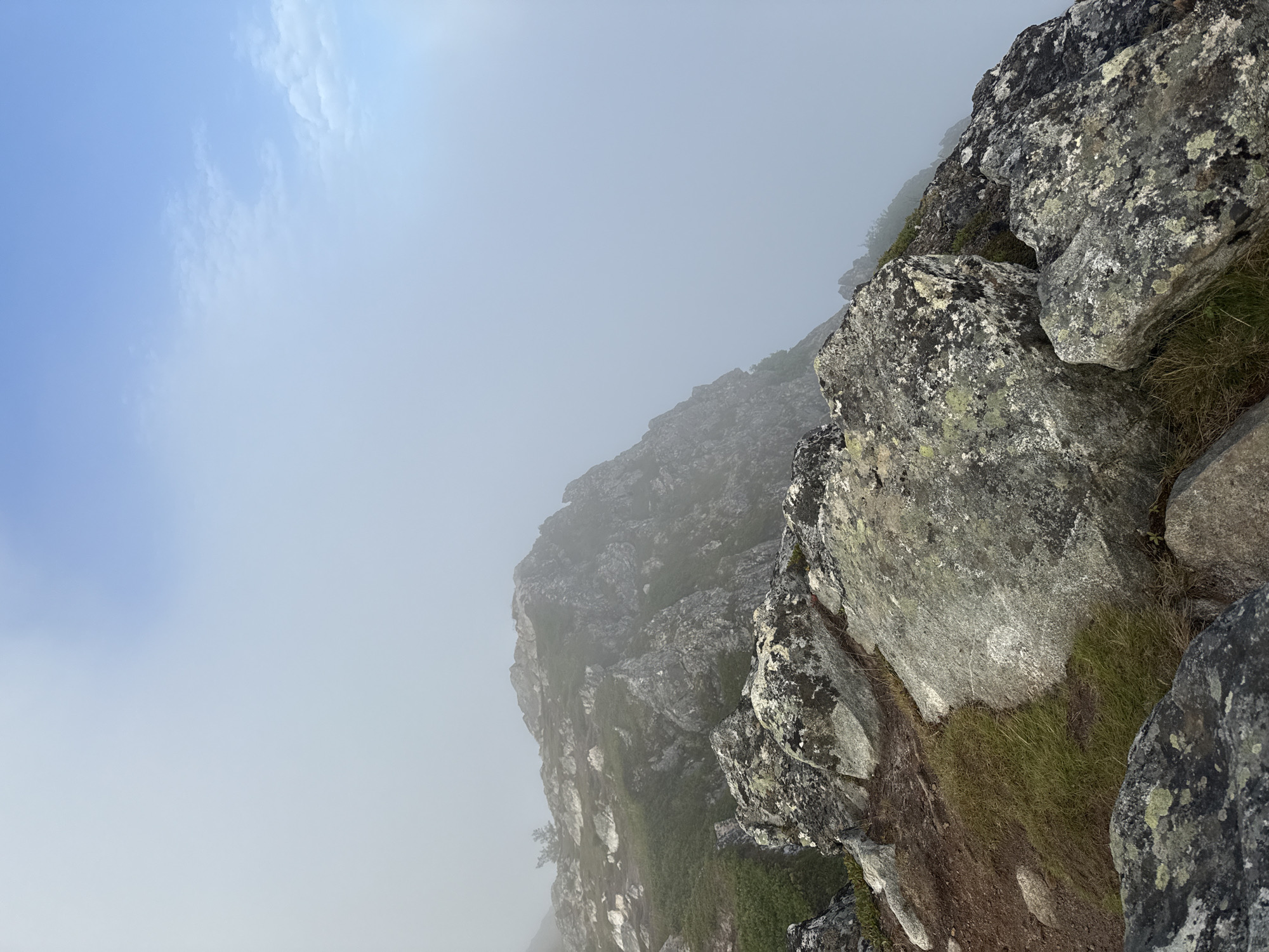 An overcast day with fog partially obscuring a mountain ridge overlooking a landscape. The foreground is dominated by large, moss-covered rocks and sparse, dry grass. A narrow, dirt path winds through the rocks on the left side of the image. In the middle ground, a rocky mountain peak emerges from the fog, with patches of green vegetation visible on its slopes. The background is filled with a thick layer of fog, with glimpses of a pale blue sky and wispy clouds visible in the upper right corner. The scene appears to be a natural, rugged terrain, likely a hiking trail or mountain summit. The location is on the summit of Trollskjærberget in Harstad, Norway.