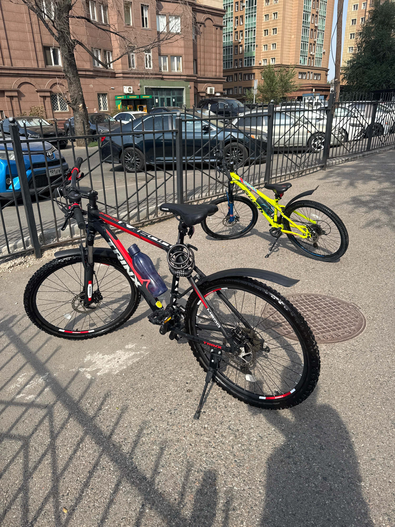 This image captures a street scene in Almaty, Kazakhstan, on a sunny day. In the foreground, a black and red Trinx mountain bike is parked on a paved sidewalk, casting a shadow. The bike features a blue water bottle attached to its frame and a combination lock secured to the seat post. Behind it, a bright yellow mountain bike is also parked. The bikes are positioned near a black metal fence that separates the sidewalk from a street where several cars are parked. In the background, a brown building with numerous windows stands alongside modern, light-colored residential buildings. The overall environment suggests an urban setting with typical city infrastructure and vehicles.