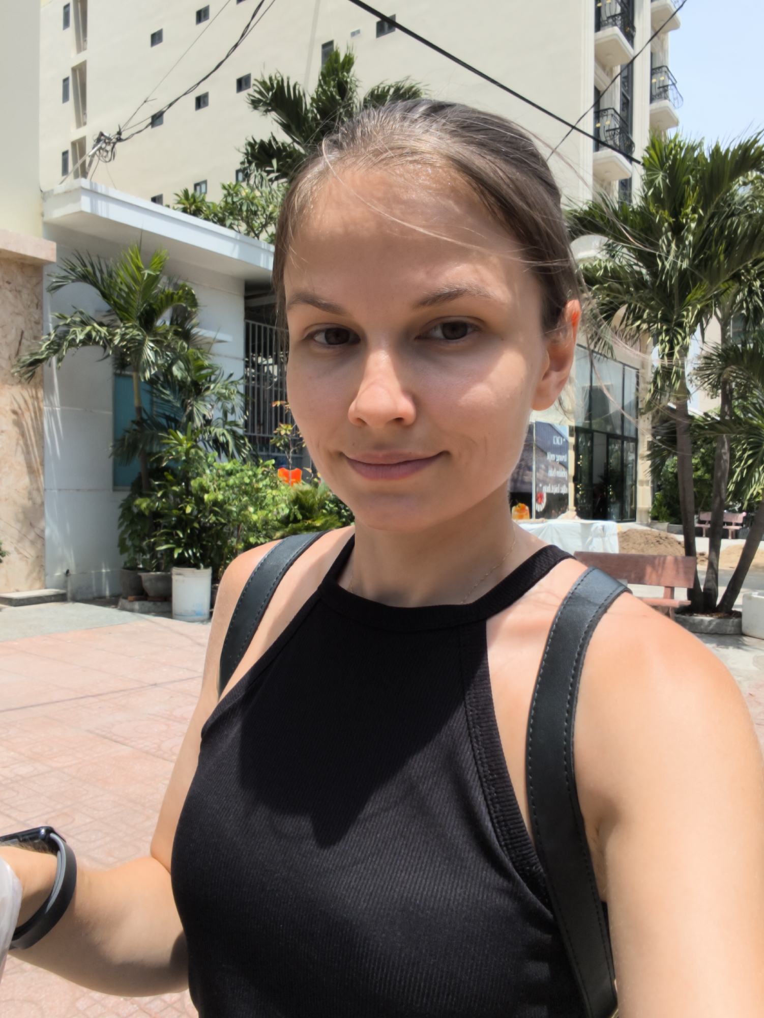 The media file shows a young woman taking a selfie outdoors. She is wearing a black tank top and a backpack. The background consists of a modern building with balconies and palm trees, suggesting a warm, sunny climate. The ground is paved with reddish-brown tiles. The time of day appears to be daytime due to the bright sunlight casting shadows.
The woman's expression is relaxed and pleasant. Her hair is pulled back, and she is looking directly at the camera. Visible on her left wrist is a black smartwatch. A white plastic bag is also partially visible in her left hand.
In the background, there is a white building with multiple floors and dark windows. Green foliage, including palm trees and other lush plants, surrounds the building. Some of the plants are in pots. There are also some construction materials and benches visible. One sign in the background has text, but it is not legible in this image. The overall setting suggests a tropical or sub-tropical urban environment, consistent with the provided location context of Bac Nha Trang Ward, Vietnam. The weather appears to be clear and sunny.