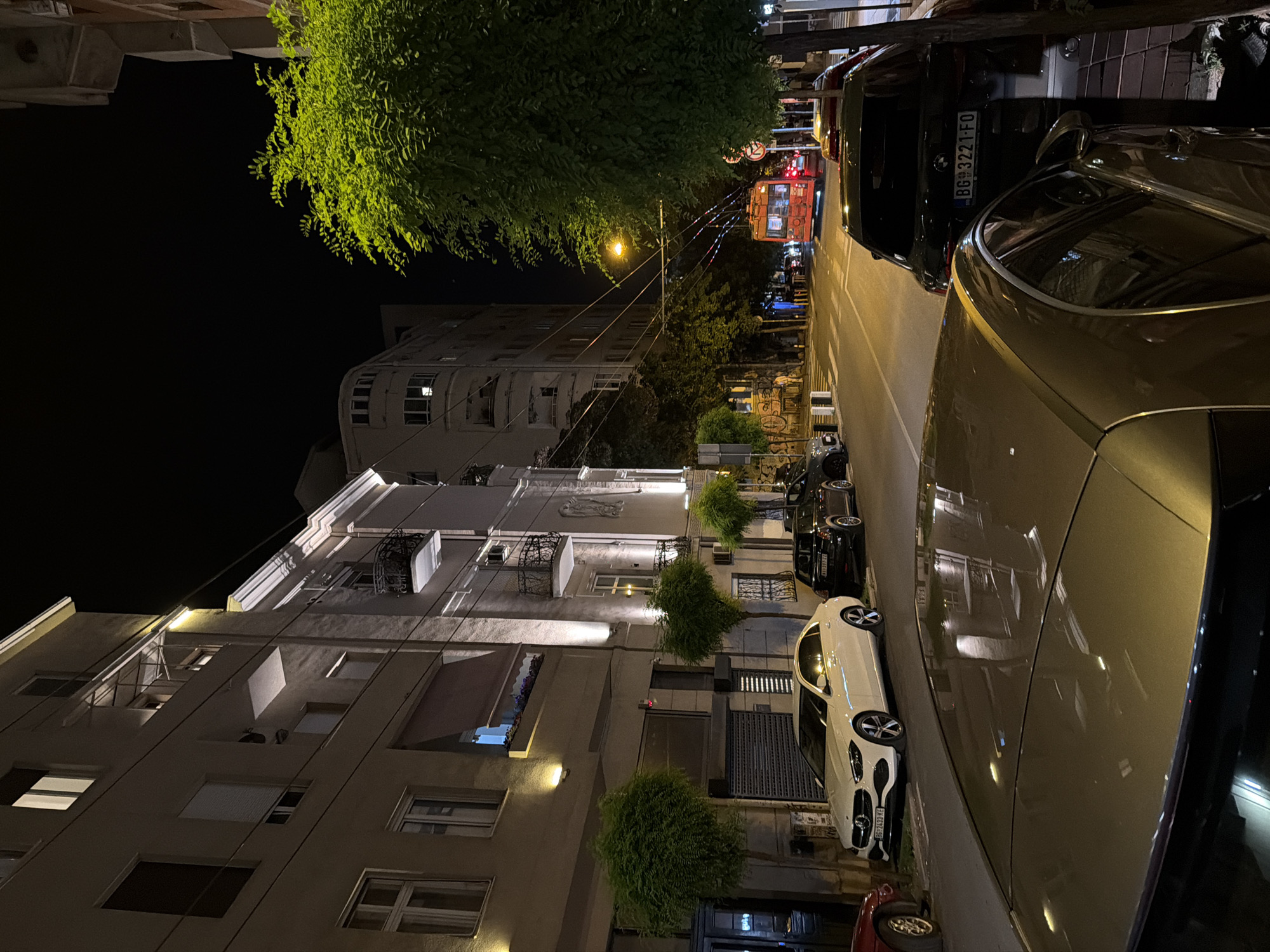 The image depicts a nighttime street scene in Belgrade, Serbia. In the foreground, the metallic surface of a car's roof is visible, reflecting the streetlights and surrounding buildings. To the left, a white Mercedes-Benz sedan is parked parallel to the curb, its headlights on. Behind it, a black car is also parked, with a license plate reading "BG 3221-FO". The street is lined with trees that are illuminated by streetlights, casting shadows on the pavement. Buildings with lit windows and balconies line the street on the left side. In the distance, an orange trolleybus is visible on the street, surrounded by traffic. The overall atmosphere is that of a quiet urban street at night.