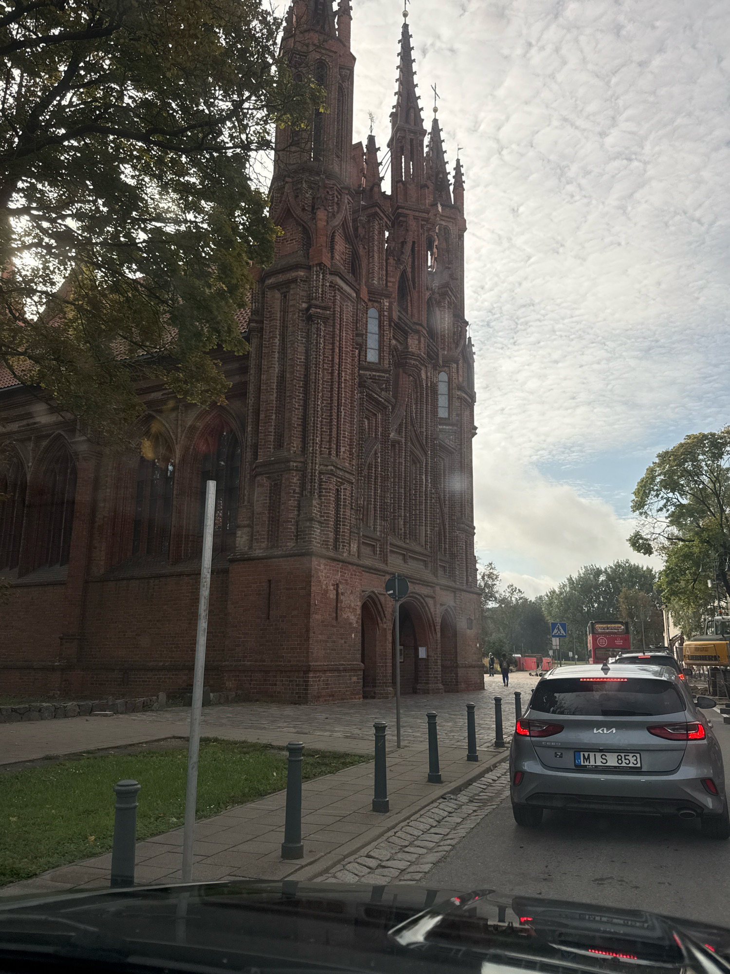 The image, taken from a vehicle, shows a view of the Church of St. Anne in Vilnius, Lithuania. The gothic brick church dominates the left side of the frame, with its characteristic spires and intricate facade. A large tree with green foliage is partially obscuring the church on the left. In the foreground, the hood of the vehicle from which the photo is taken is visible, reflecting the sky. A silver Kia Ceed car with the license plate "MIS 853" is in the right lane of the street, facing away from the viewer and stopped behind other vehicles. A double-decker tour bus and construction equipment are also visible further down the street. The sky is overcast with scattered clouds. The street appears to be under some form of renovation or construction, with orange barriers visible on the right side of the road. People are walking on the sidewalk next to the church.