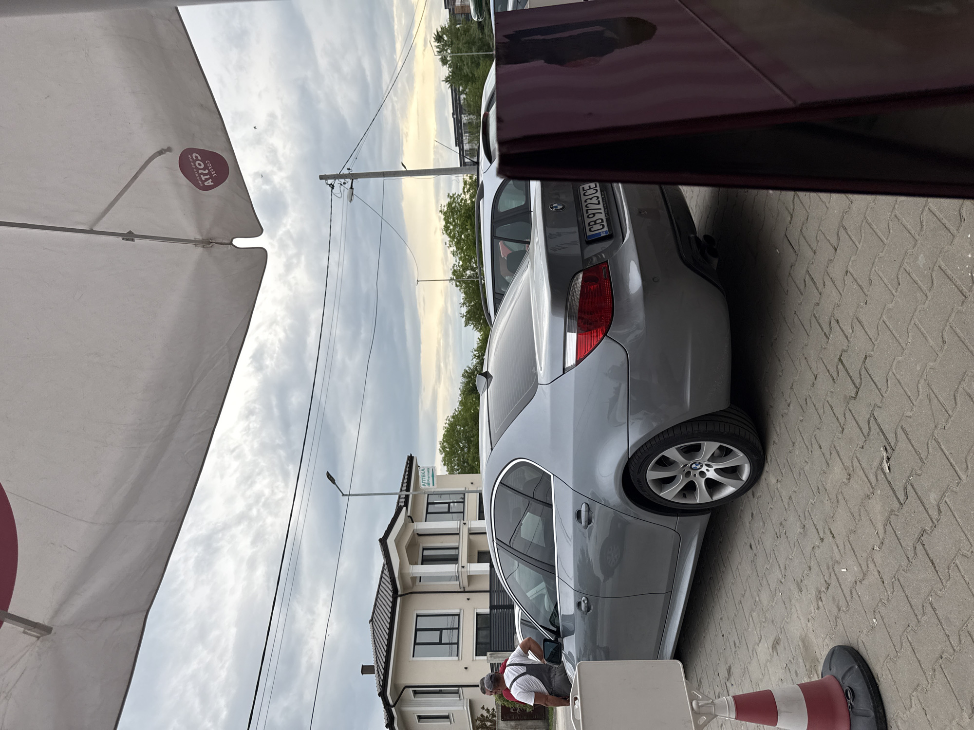 The image depicts a grey BMW car parked on a paved area, likely a sidewalk, in an urban setting. Part of a white sun umbrella with a "COFFEE ATOS" logo is visible in the upper left, suggesting a cafe or outdoor seating area. A building with white facade and dark windows is in the background. A man in a grey overall and a white shirt appears to be working on the car. Overhead power lines are visible against a cloudy sky. The license plate of the car is "CB 9723 CE". Based on the license plate, this photo was taken in the city of Plovdiv, Bulgaria.