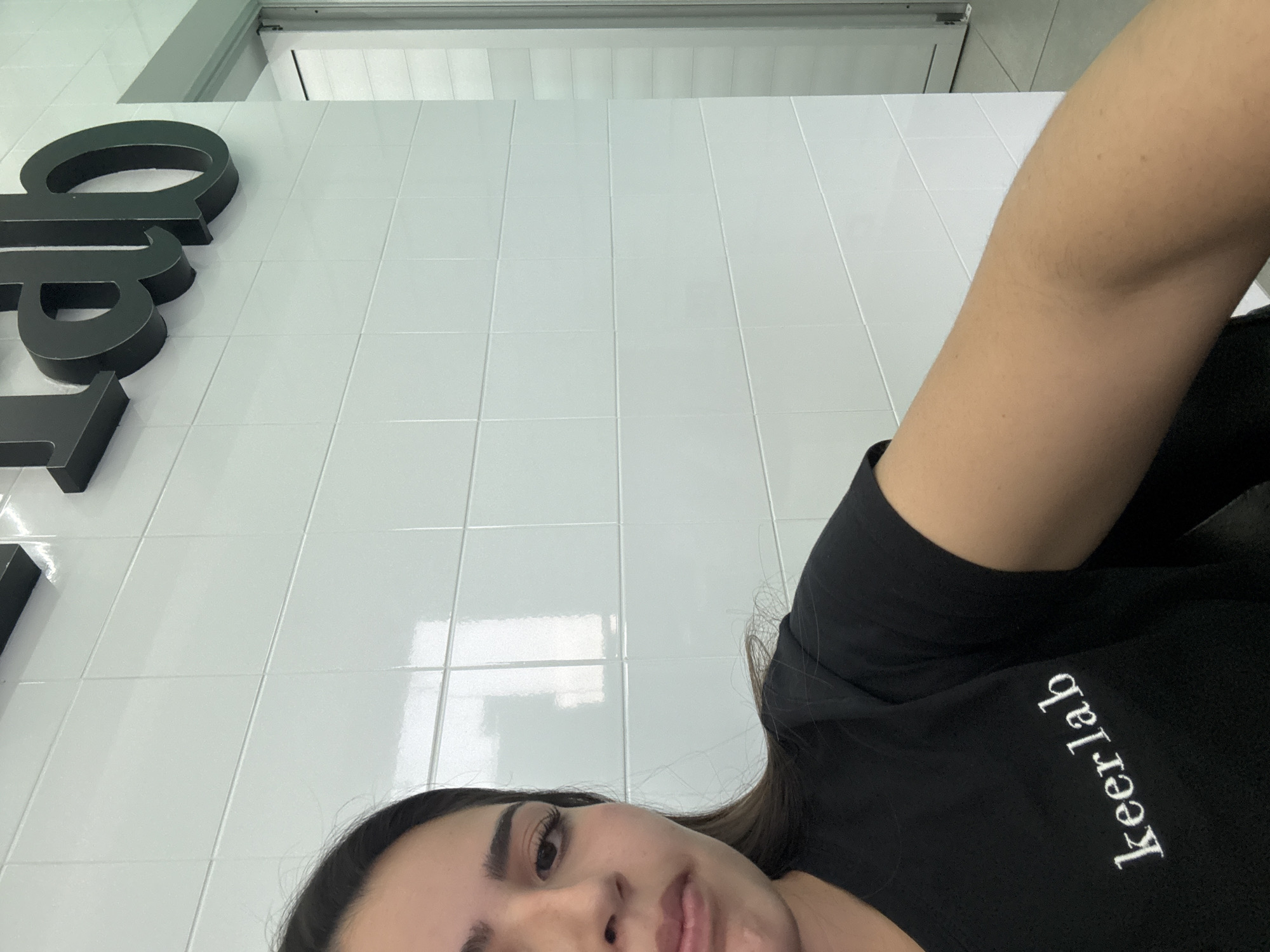 The image captures a close-up, angled perspective of a person indoors. The subject, a female with dark hair and eyebrows, occupies the lower right portion of the frame, with her face partially visible, looking towards the camera. Her right arm is extended across the mid-right. She wears a black t-shirt with white text that reads "keer lab," which appears reversed due to the camera's orientation.
The setting appears to be a modern, clean interior, likely a laboratory or commercial space, possibly in San Bernardino Tlaxcalancingo, Mexico. The background features a pristine white tiled wall with subtle reflections of light and the environment. Prominently displayed on the wall are large, dark, three-dimensional letters. Due to the extreme angle and potential reflection, the visible letters appear as "qap" or "gap" when read from left to right, but they are clearly part of a larger sign, highly likely "Keer Lab" given the matching text on the subject's shirt. Above the tiled wall, a light-colored ceiling or upper wall section with a grey frame and a white panel is visible.
The overall impression is a casual, possibly playful, moment captured indoors. The lighting is bright and even, suggesting daytime. The tilted nature of the shot creates a dynamic and unconventional portrait.