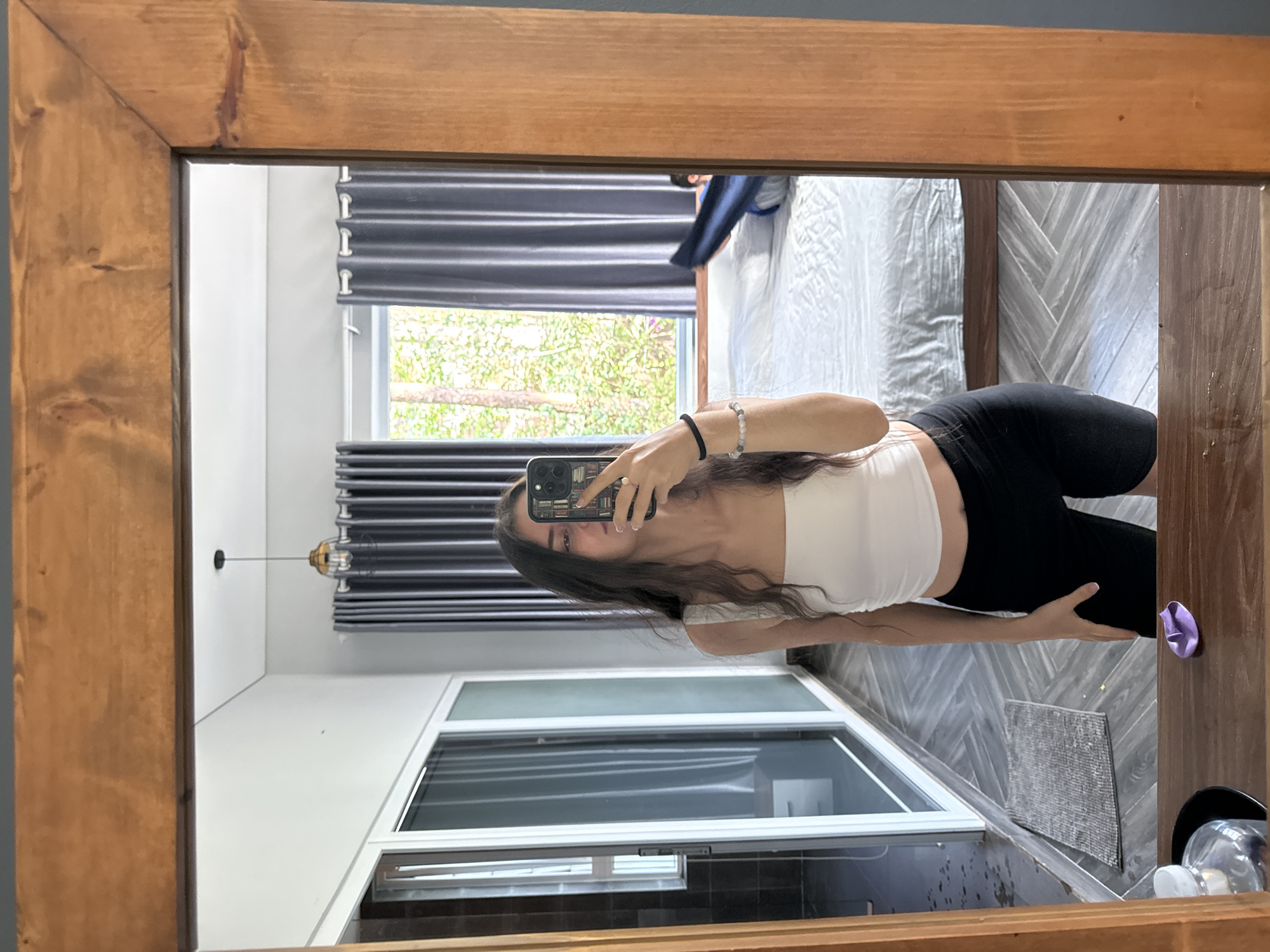 A young woman is visible in the reflection of a wooden-framed mirror, likely taking a photo of herself with her phone. She is wearing a white crop top and black leggings. The background includes a window with gray blinds, a view of foliage outside, and a bed with gray and white bedding. The room appears to be in Da Lat, Vietnam, as suggested by typical architectural style and foliage. The floor is decorated with gray tile laid in a geometric pattern. A window is visible in the bottom left corner of the reflection. A purple object rests on the edge of the mirror.