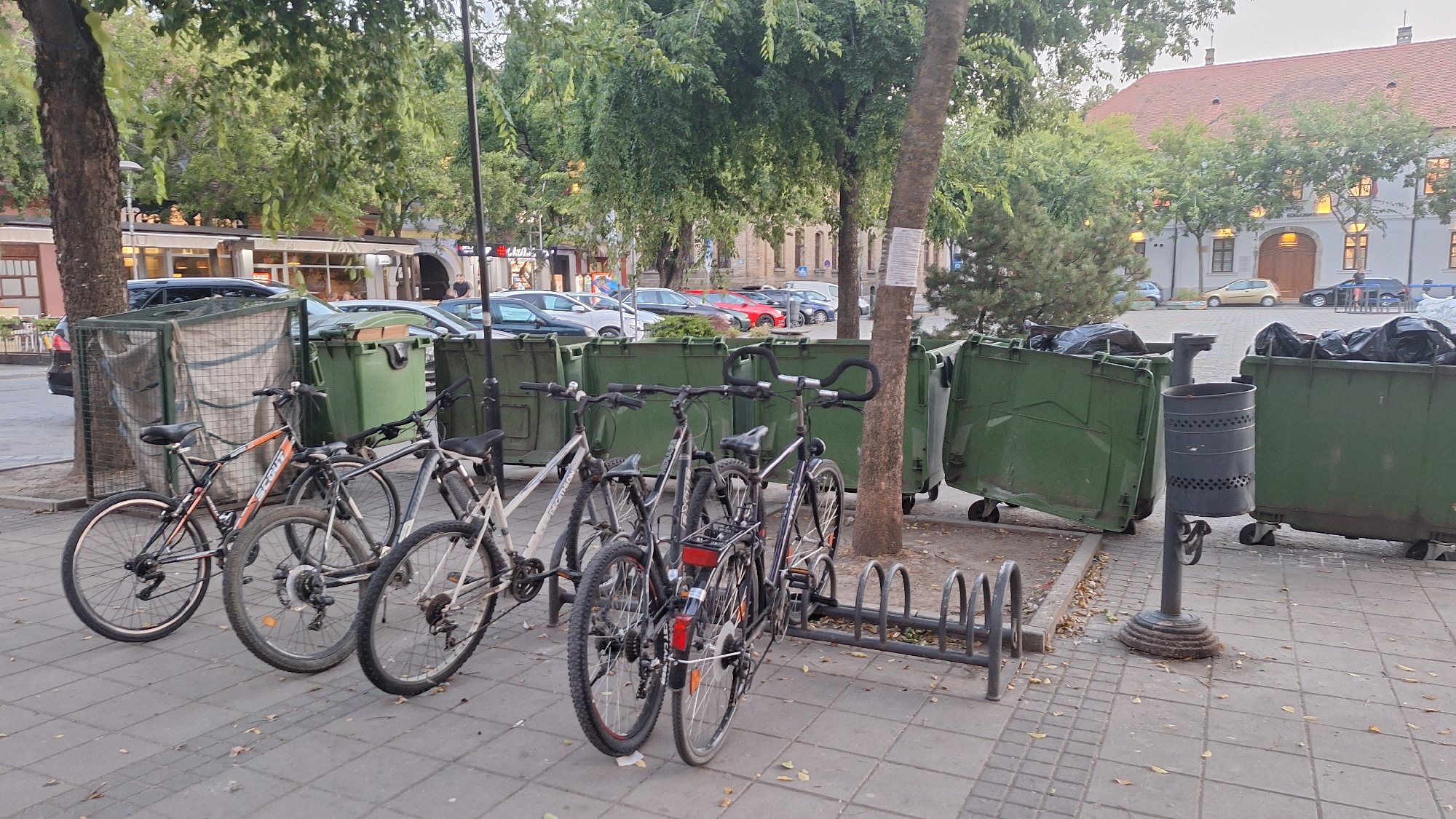 The image depicts a street scene in the city of Sombor, Serbia, likely in front of commercial establishments. In the foreground, several bicycles are parked at a bicycle rack. These include an orange and black bicycle, a silver bicycle, and a dark-colored bicycle. Behind the bicycles, large green garbage bins are visible, some with black trash bags inside. A smaller, grey trash can is mounted on a pole to the right of the green bins. The ground is paved with square tiles. In the background, trees with lush green foliage line the street. Further back, buildings with windows and a red-tiled roof are visible, suggesting an urban setting. Cars are parked along the street, and some people are seen walking in the distance. The lighting suggests it is daytime, possibly late afternoon or early evening.