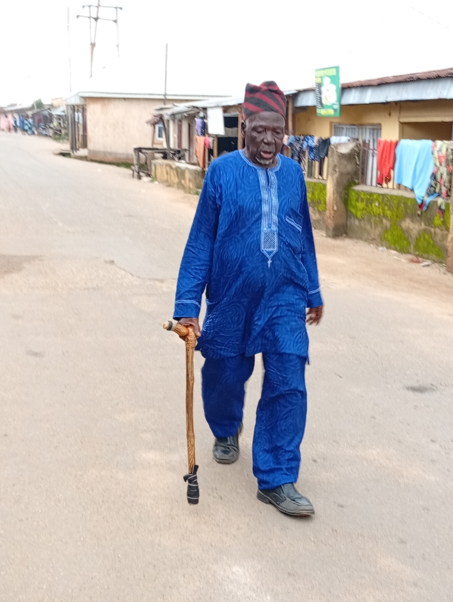 An elderly Black man wearing a blue traditional outfit and a striped cap walks with a wooden cane on a paved street in a residential area of Jos, Nigeria. The man's outfit consists of a long-sleeved top with intricate blue embroidery on the chest and matching loose-fitting trousers. He holds a rustic wooden cane with a carved handle in his right hand. He is walking towards the right side of the frame.
The environment appears to be a street lined with modest buildings and open stalls. Clothes are hung on a line in front of one of the buildings, suggesting a residential or commercial area. A sign with green lettering is visible above one of the stalls. The sky is overcast, and the lighting suggests it might be late afternoon or early morning. The street surface is asphalt and shows signs of wear. The overall impression is of a typical street scene in a developing urban area.