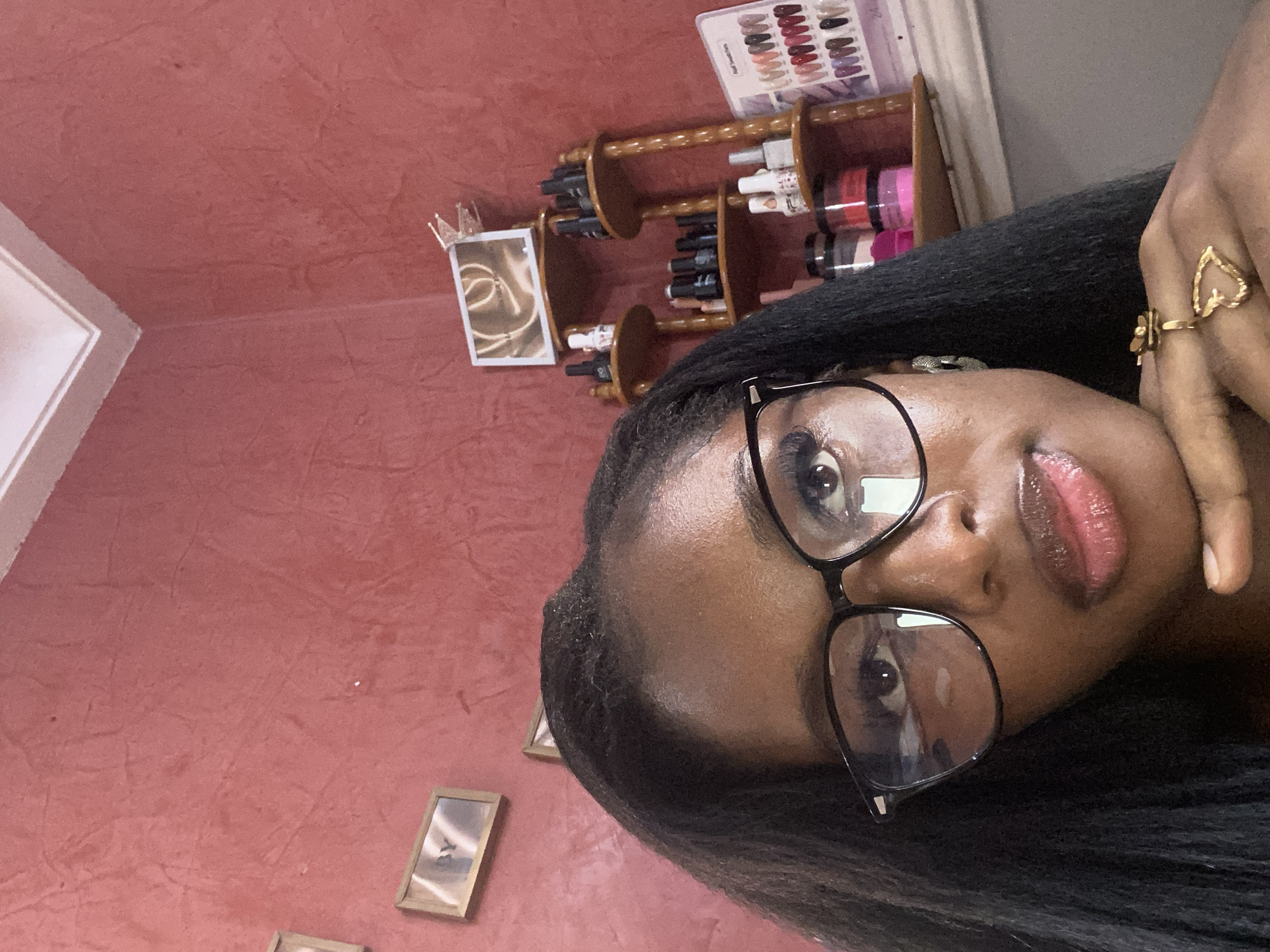 A woman with dark skin and long, dark hair is taking a selfie. She is wearing black-rimmed glasses and has her hand resting on her chin. She is also wearing a gold ring on her right hand. In the background, there is a red, textured wall with two framed pictures. To the right, there is a wooden shelf with various nail polish bottles and a nail swatch chart. The scene appears to be indoors, possibly in a home or salon setting. Based on the visual cues, such as the nail products and the overall aesthetic, the location is likely a beauty salon or a personal grooming space within a residential area in the city of Jos, Nigeria.
