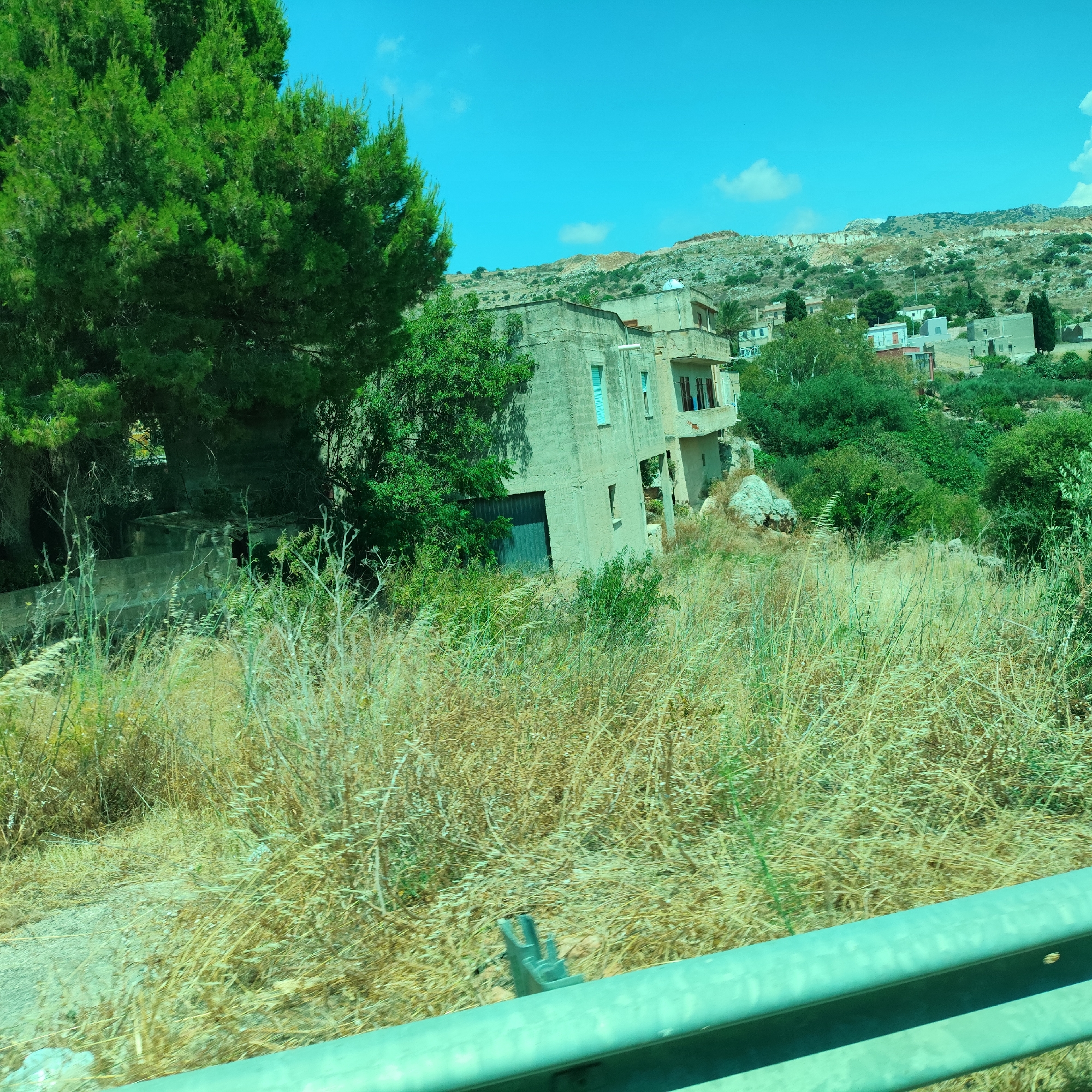 The image depicts a rural, hilly landscape under a bright blue sky with scattered clouds. In the foreground, dry, golden grass and various green plants grow wild. A light blue metal guardrail runs horizontally across the bottom of the frame.
Dominating the middle ground is a light-colored, weathered building constructed from what appears to be concrete blocks. This structure has multiple levels and balconies, with some windows boarded up and others showing dark interiors or balconies with visible windows. A dark garage-like door is visible on the lower level of the building. To the left of this building, a large, lush green evergreen tree with dense foliage stands prominently.
Behind the main building, the landscape continues to ascend with more greenery and other smaller buildings scattered among the hills. The overall impression is one of a somewhat neglected or rustic setting, possibly in a Mediterranean or arid region, given the dry grass and the style of the architecture. The bright sunlight suggests it is daytime, likely midday or early afternoon. There is no discernible activity or people visible in the image. No text is present.