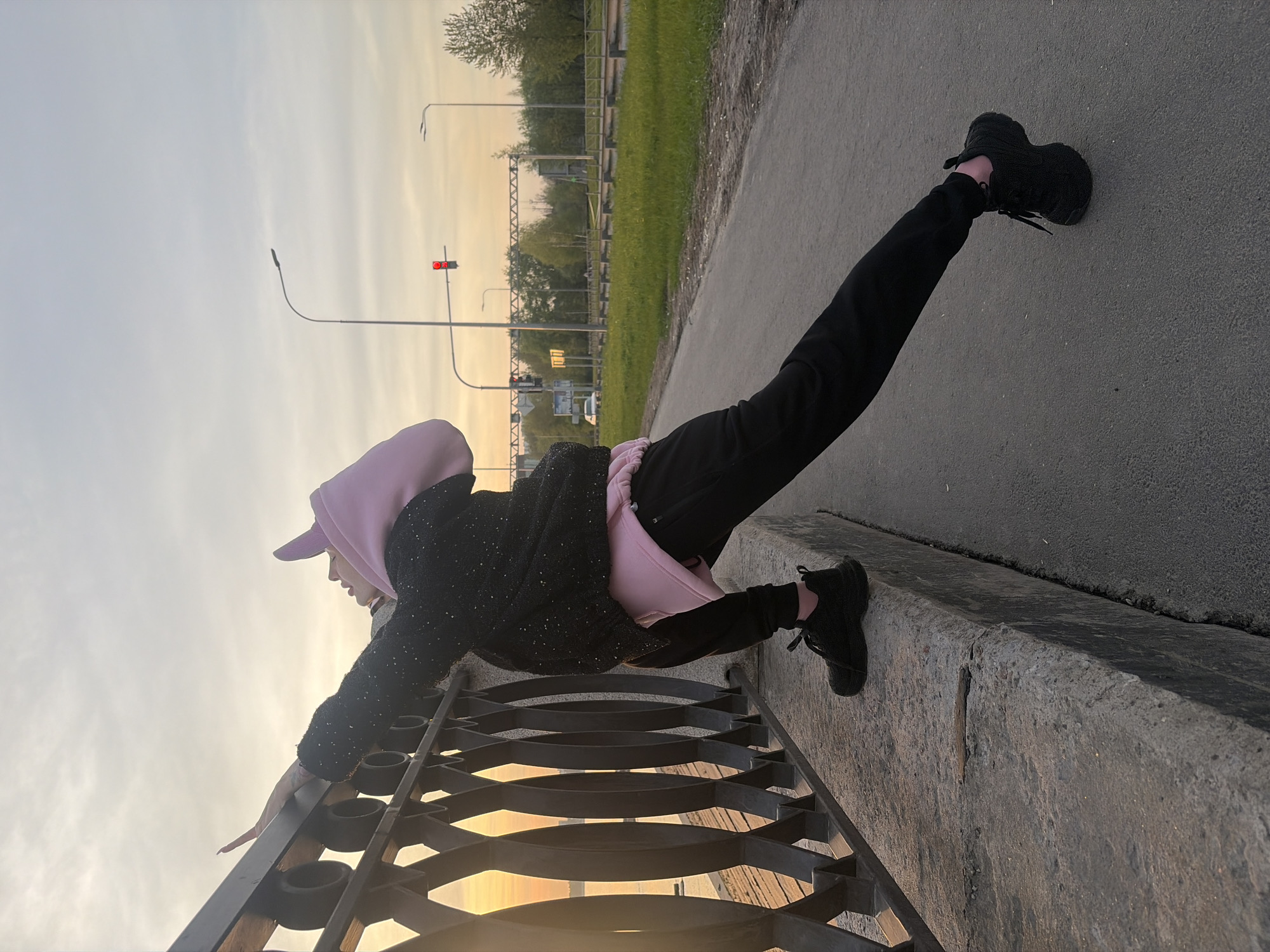 A sideways, wide shot depicts a person doing a plank on a concrete ledge in what appears to be Saint Petersburg, Russia. The person is wearing black pants, a black jacket with speckles, a pink hoodie, and black sneakers. They are leaning on a black, decorative metal fence with a sunset visible behind it. Their feet are resting on the concrete ledge. In the background, there is a street with a traffic light, trees, and streetlights. The sky is a mix of light blue and orange, indicating it is either sunrise or sunset.