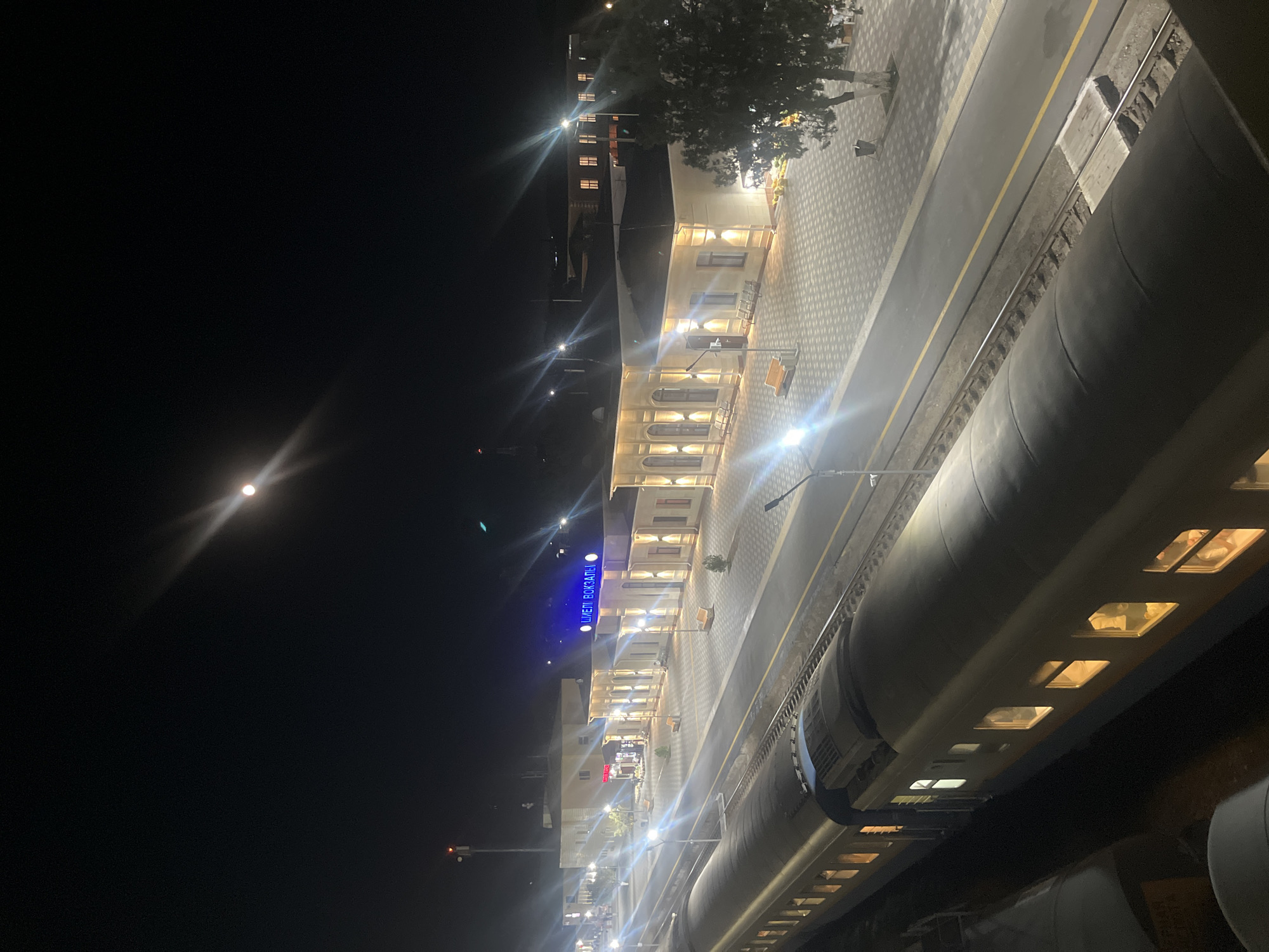 The image depicts a night scene at the railway station in Şïeli, Kazakhstan. A train is parked on the tracks, with its illuminated windows suggesting occupancy. The station building is well-lit, featuring a prominent sign with blue illuminated lettering that reads "ШІЕЛІ ВОКЗАЛЫ" (which translates to "Shiely Station" in Kazakh). The station platform is paved with patterned tiles and lined with benches. Streetlights illuminate the area, and a bright moon is visible in the dark sky, with a lens flare effect. A lone tree stands to the right of the station building, and a trash receptacle is visible near its base. The overall environment appears clean and orderly.