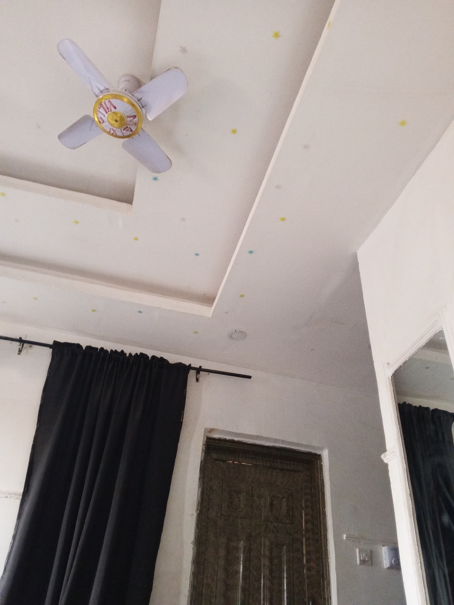 The image shows a room interior. A white ceiling fan with gold and red details is mounted on a white ceiling. The ceiling has recessed sections and is decorated with small star-shaped stickers of yellow and blue colors. Below, a black curtain hangs on a black rod, partially obscuring a dark brown wooden door with a raised panel design. A white wall and a white-framed mirror are visible to the right. Electrical outlets are present on the wall near the door. The location is Baganakwo, Nigeria.