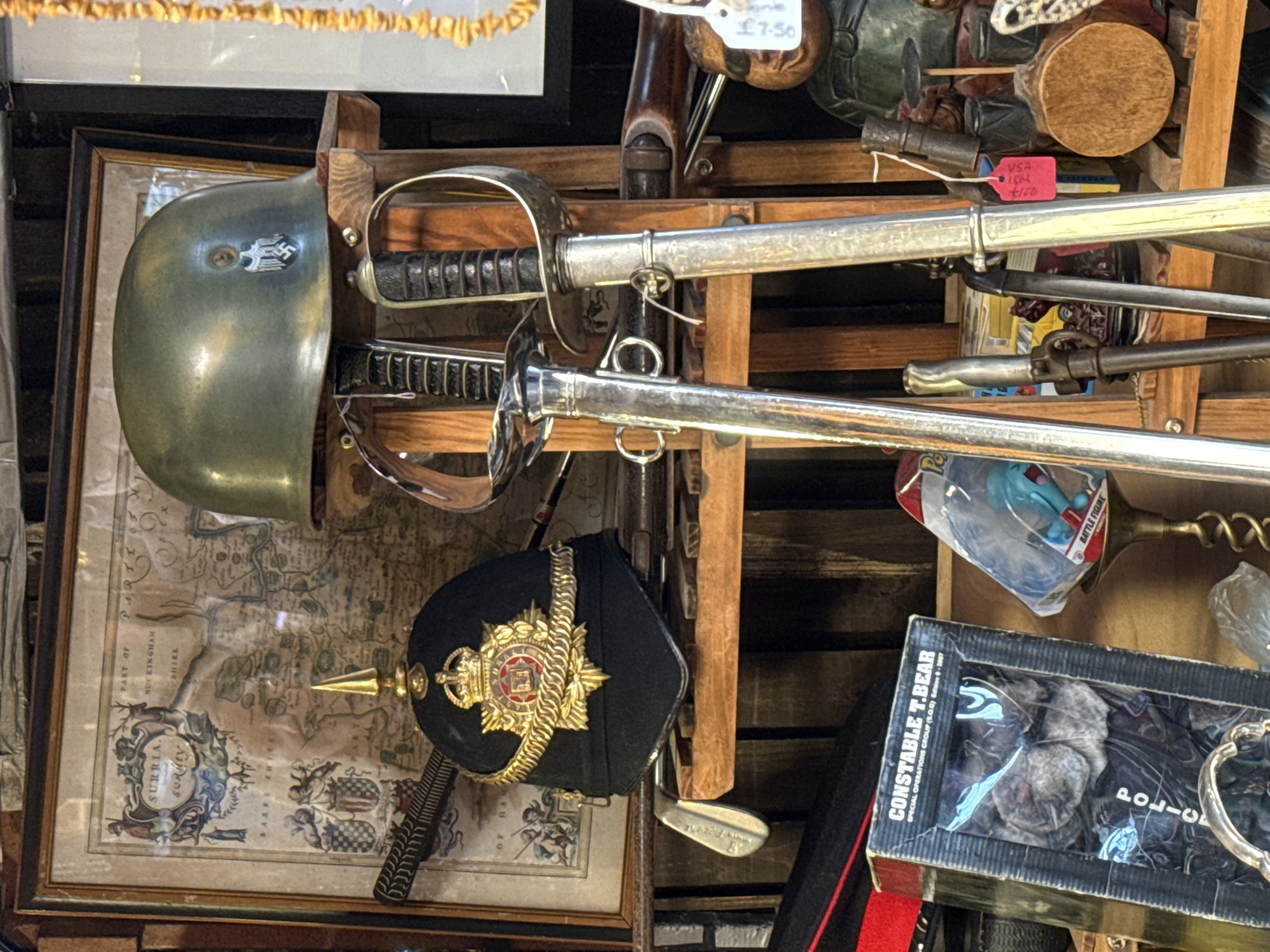 The image depicts a shelf in a shop, cluttered with various vintage and antique items. Two swords with silver hilts are prominently displayed, along with a dull green helmet. To the left, a framed vintage map labeled "SURRIA, SURMEY" is visible. Below the helmet, a black military hat adorned with gold braid and a central crest sits beside the map. A box labeled "CONSTABLE T.BEAR" is partially visible in the lower right corner. Additional items, including figurines, wooden objects, and small weapons, are scattered on the shelf. A price tag reading "£7-50" and another with "VSA 1864 $100" suggest the items are for sale. The overall impression is of a cluttered antique shop or market stall, potentially located in London, United Kingdom given the style of the items and the British pound price tag.