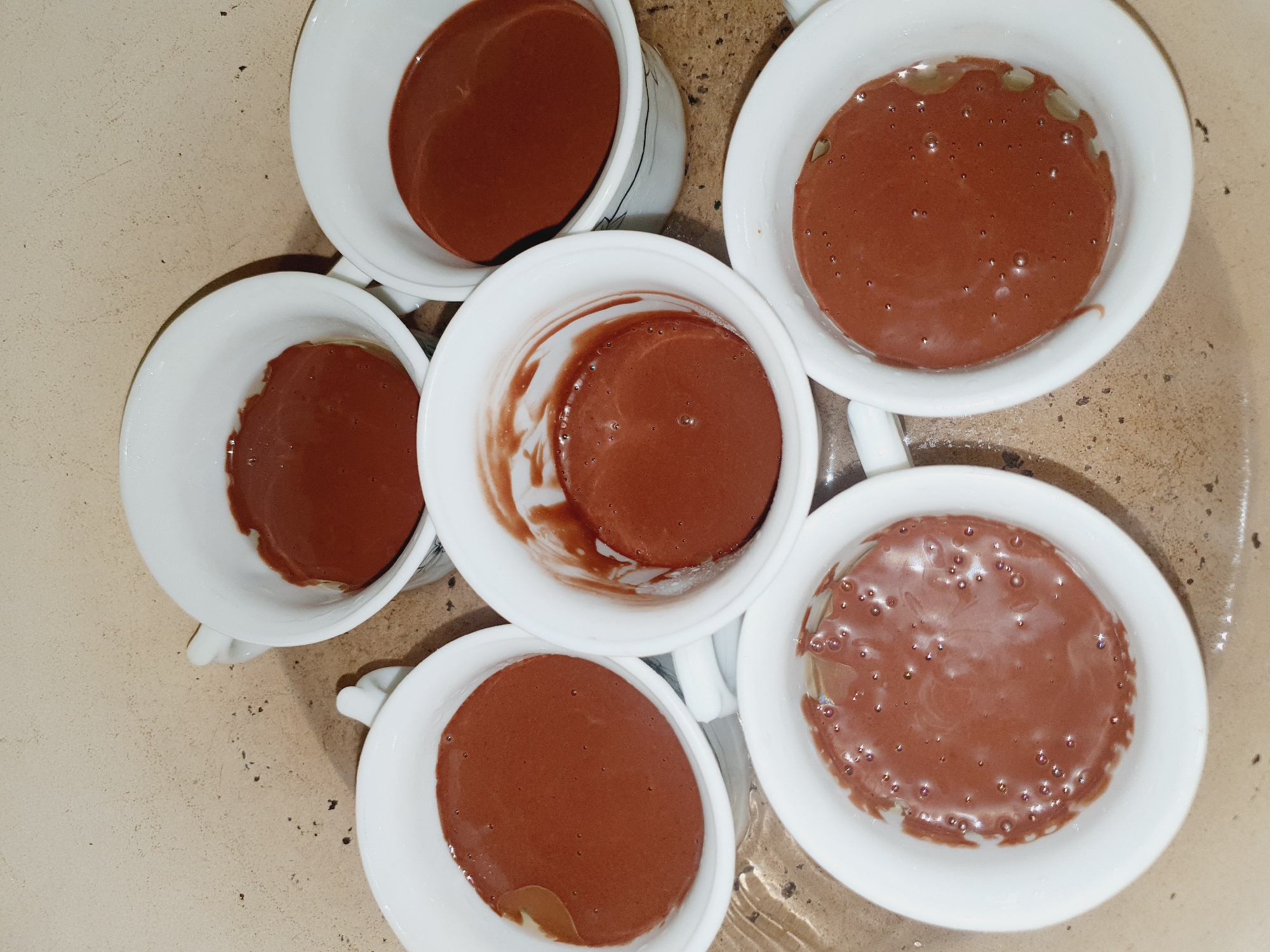 The image shows five white ceramic cups filled with a brown liquid, likely chocolate pudding or mousse. The cups are arranged in a circular pattern on a speckled, beige surface that appears to be a countertop. Some cups have small bubbles on the surface of the liquid, suggesting it may have been recently prepared or poured. The lighting is overhead, casting subtle shadows. There are no people or discernable landmarks visible that would indicate a specific location in Shiraz, Iran. The scene is a close-up, overhead shot of the dessert preparations.