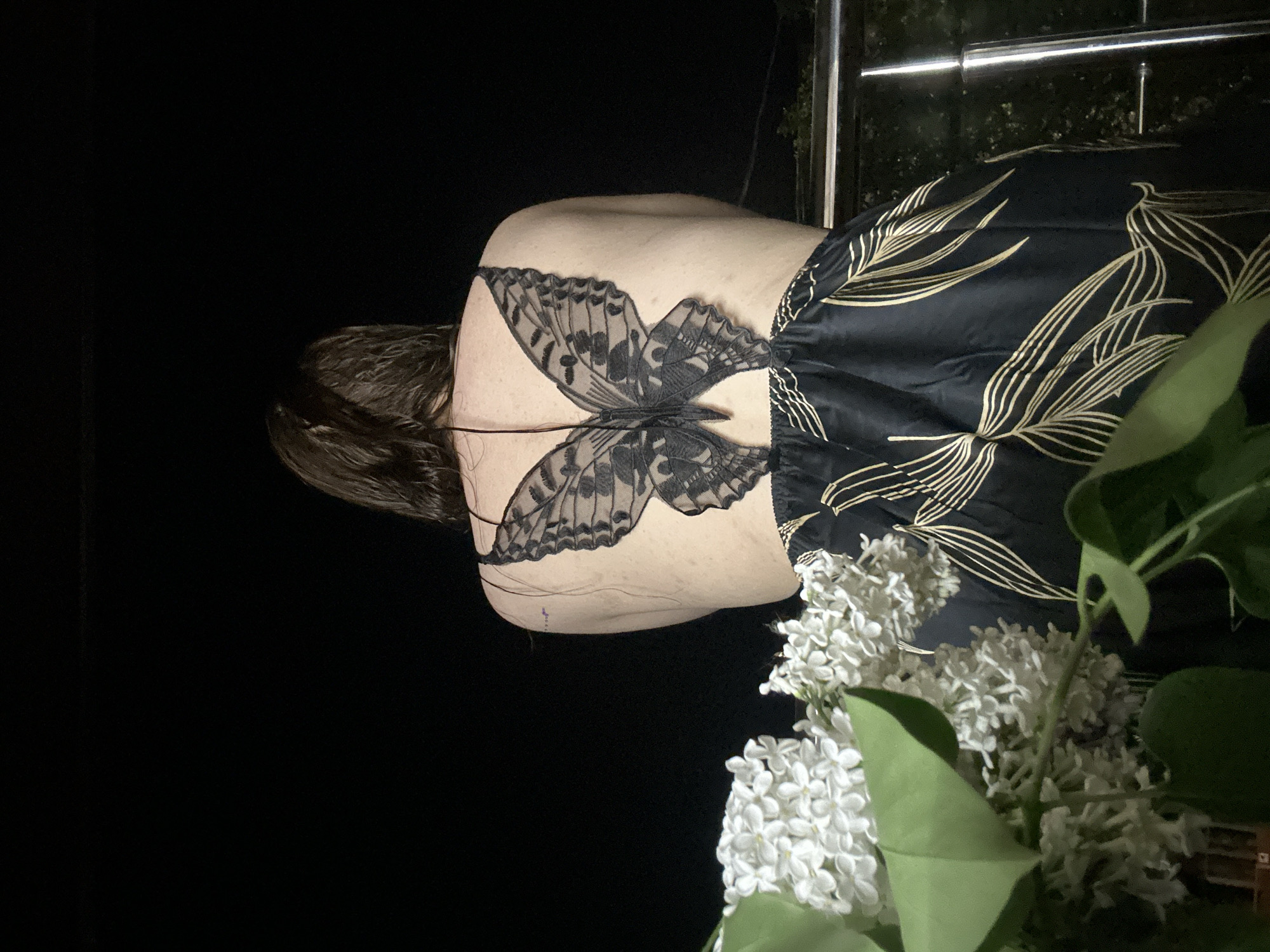 A person with a butterfly tattoo or design on their back is standing on a balcony in Nizhny Novgorod, Russia. They are wearing a dark garment with gold leaf patterns. White lilac blossoms are visible in the foreground. A silver railing can be seen in the background. The lighting suggests it is evening or night.