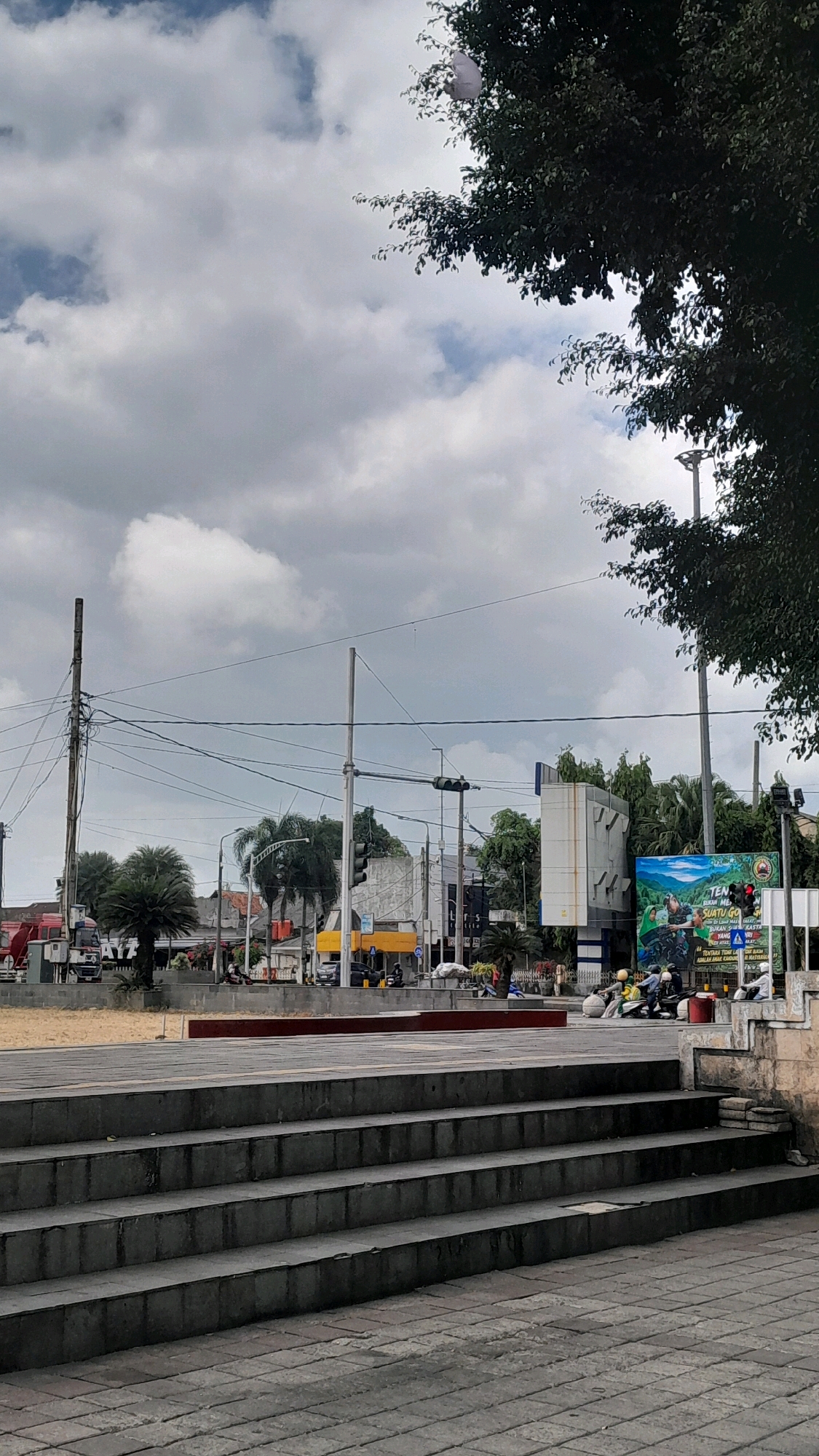 The image depicts an outdoor scene in the city of Cilacap, Indonesia, likely a public square or intersection. In the foreground, a set of wide, stone steps leads up from a paved area. Beyond the steps, a sandy area is visible, bordered by a low red wall. Further back, a street is active with traffic, including several motorcycles and a few cars. Buildings line the street, some with visible signage. A large billboard with a colorful image and text in Indonesian is prominent on the right side of the street. Overhead, a network of utility poles and wires crisscrosses the sky. Trees, including palm trees and a large tree in the upper right corner, are scattered throughout the scene. The sky is filled with dramatic, dark clouds, suggesting an overcast or approaching storm. A white object, possibly a plastic bag or balloon, is snagged in the branches of the tree in the upper right. The overall impression is of a busy urban environment under a dynamic sky.