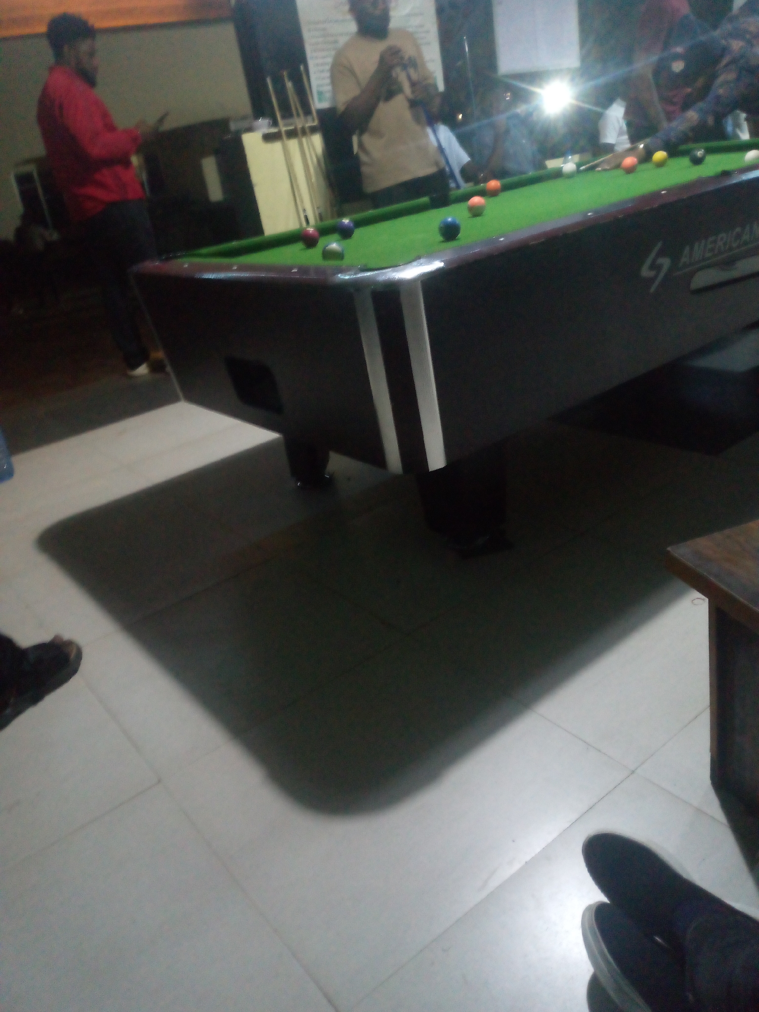 The image depicts an indoor scene, likely a bar or recreation center, featuring a pool table. The lighting is dim, with a bright light source appearing to be a flash or overhead lamp creating strong shadows. Several individuals are visible, some in focus and others blurred. In the foreground left, a person in a red shirt and dark pants is standing, looking down at their phone. On the pool table, several pool balls are scattered across the green felt surface. The table itself is dark with white trim. In the background, other individuals are present, some gathered around the table. One person appears to be in the process of taking a shot with a pool cue. The floor is tiled. The image does not contain any identifiable landmarks or cues that would definitively place it in Jos, Nigeria.