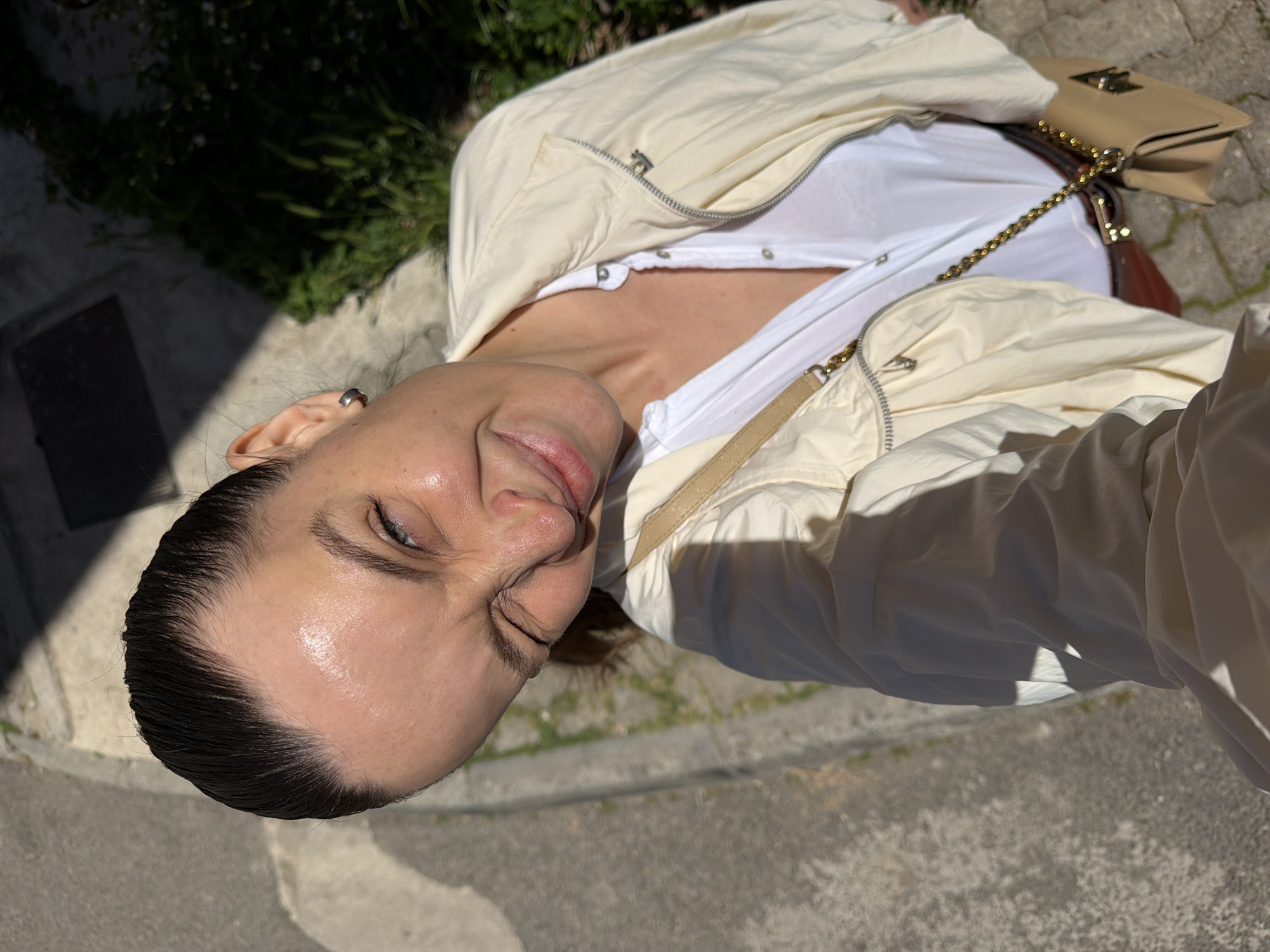 A downward-angled photograph depicts a woman in an outdoor setting. She wears a white t-shirt, an unzipped light-colored jacket, and has dark hair pulled back from her face. A tan-colored purse with a gold chain strap is visible. The background includes foliage and a paved area, likely a sidewalk. Based on the architectural style, vegetation, and lighting, the location could plausibly be in or near the city of Budva, Montenegro.